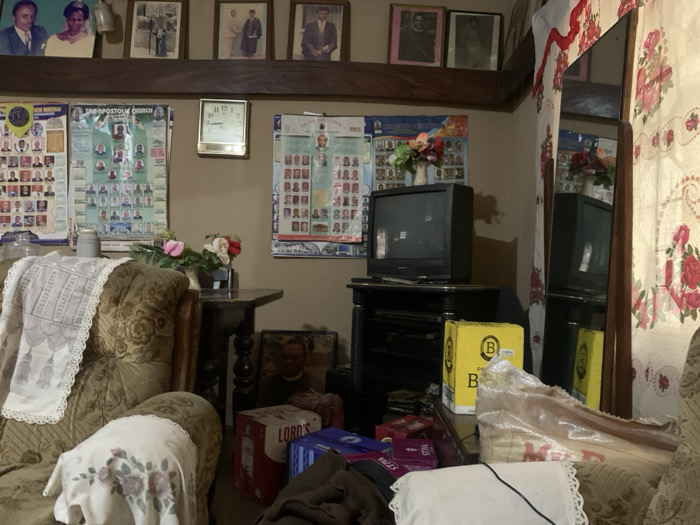 This image depicts a cluttered indoor residential living space located in Uyo, Nigeria. The upper portion of a beige wall features a dark wooden beam shelf supporting six framed photographs, predominantly portraits of individuals or couples. Below the shelf, three large laminated posters are affixed to the wall; the visible text indicates "THE APOSTOLIC CHURCH," and each poster displays a grid of small portrait photos and names, likely membership or leadership listings. A golden-framed wall clock is positioned between the left two posters.
In the left foreground, a heavily upholstered armchair with a brown and gold floral pattern is draped with white, lace-edged fabric coverings. To its right, a small dark wooden side table holds a vase containing artificial pink and white flowers. Towards the center-right, a black CRT television set rests on a dark entertainment stand. A large, dark-framed mirror stands vertically against the wall on the far right, reflecting a floral-patterned curtain with red and pink roses, and parts of the room.
The lower area of the scene, particularly near the television and mirror, contains various objects, including a prominent red and white box labeled "LORD'S," a yellow box with a black 'B' logo, and several smaller blue and purple boxes. A framed portrait leans against the wall at floor level in the lower right. No individuals are present in the scene, which appears static and unoccupied.
