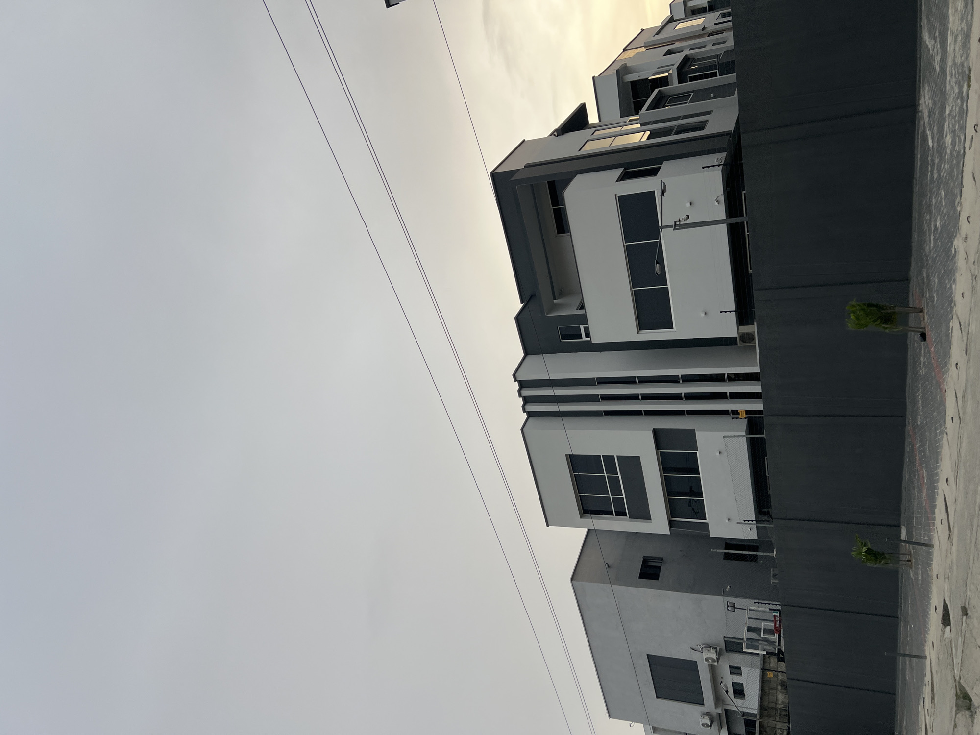 The image captures a modern urban or suburban street scene in Alaguntan, Nigeria, viewed from a low angle looking towards multi-story buildings and the sky. The sky, which dominates the left side of the frame, is hazy and light gray, subtly transitioning to a brighter, yellowish-white tone towards the upper right, suggesting late afternoon or early evening light. Several thin, dark utility lines stretch horizontally across the upper portion of the sky.
On the right side of the frame, a cluster of contemporary buildings displays a striking geometric design. They feature predominantly white and light gray facades, contrasted with dark gray or black accents, particularly in window frames and recessed architectural elements. The buildings incorporate large, dark-framed windows and what appear to be balconies or deep overhangs. An external air conditioning unit is visible on a lower section of one building.
In the immediate foreground on the right, a dark gray perimeter wall runs along the edge of what appears to be a paved or tiled ground surface, showing some texture and slight wear. A small, sparse green plant or tree sapling stands near this wall. In the background, behind one of the walls, the faint outline of a basketball hoop can be seen. The scene is devoid of people or any direct activity, presenting a static and modern environment. No visible text is present in the image.