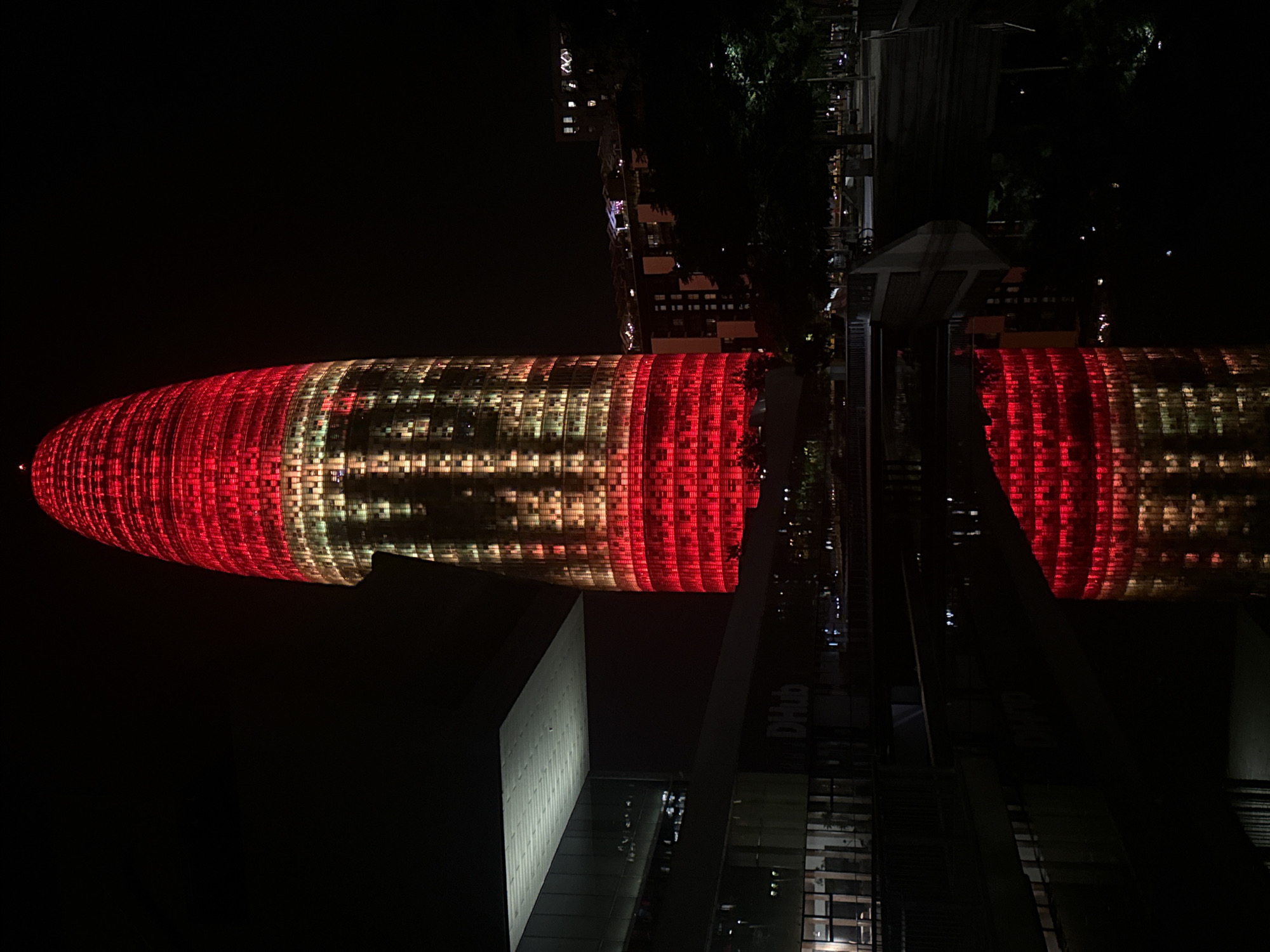 The image captures the Torre Glòries, a skyscraper located in Barcelona, Spain, at night. The building is illuminated with red and yellow lights, creating a striking visual effect against the dark sky. The lower part of the skyscraper displays the word "DHUB," indicating the Design Hub Barcelona, which is housed within the building. The skyscraper's reflection is visible in the dark water body in the foreground, doubling the illuminated structure. Adjacent to the skyscraper, a modern building with a flat roof and glass facade is partially visible. Trees and streetlights line the background, suggesting a public urban space. No people are clearly discernible in the image. The overall scene is characterized by artificial illumination and architectural prominence.