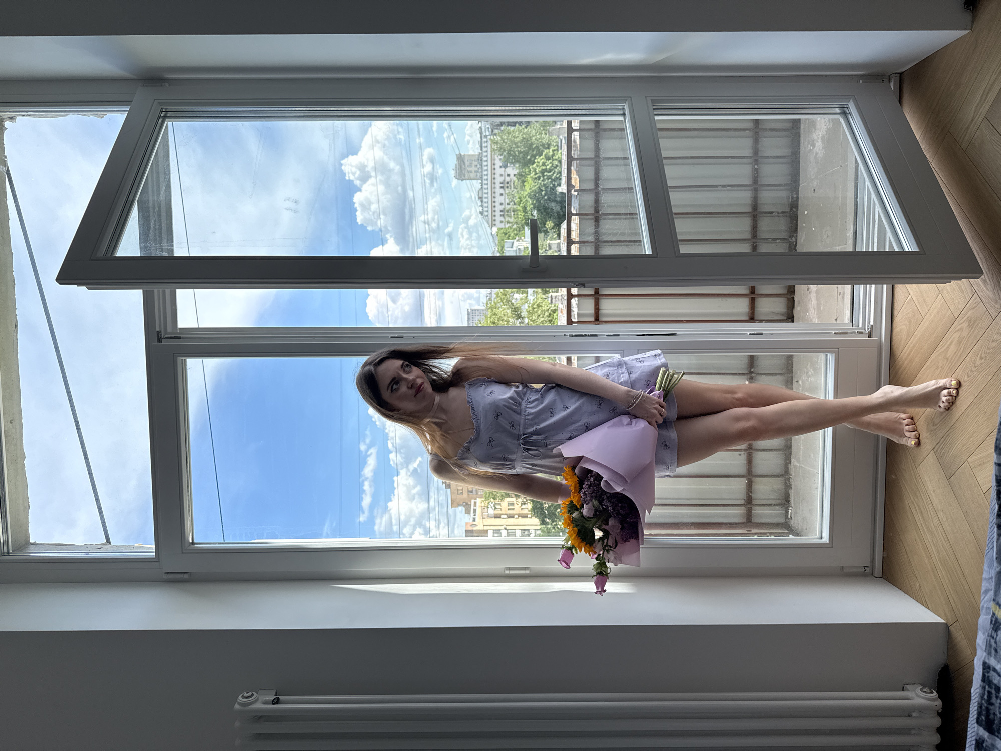 A woman is posing in an open window with a bouquet of flowers. The window frame is white. The woman is wearing a light purple dress with a pattern of dark bows. She is holding a bouquet of flowers wrapped in light purple paper. Her fingernails and toenails are painted gold. Outside of the window is a view of Moscow, Russia. The building in the background is located at Spiridonovka 24/1, Moscow, Russia.