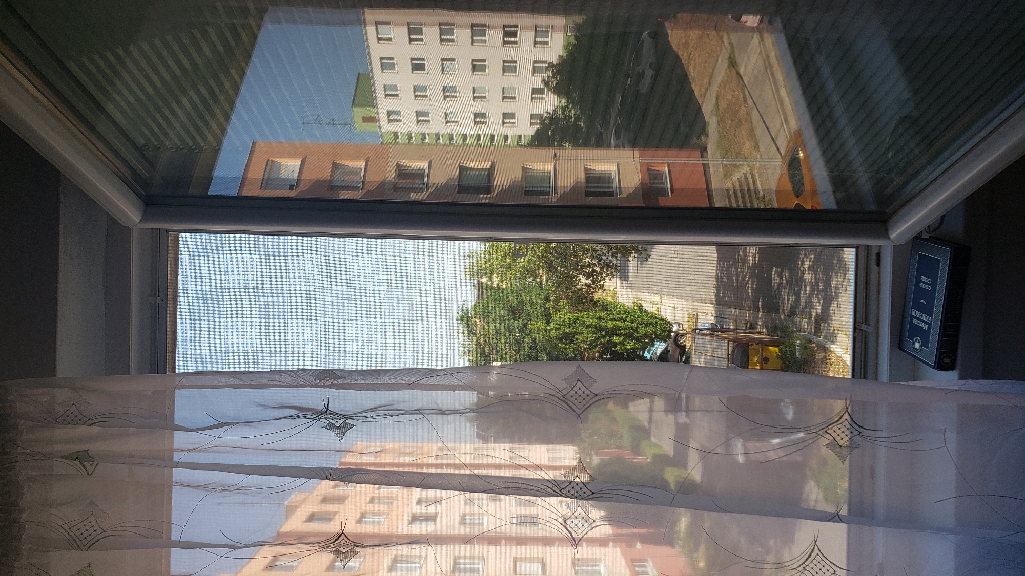 The image shows a view from a window in Bratislava, Slovakia. The window has a sheer curtain on the left side and a window screen. Outside the window, there are apartment buildings, trees, and a street. In the foreground, on the windowsill, there is a black book with a blue and white cover. The book's title is visible and reads "Margaret Mitchell: Gone With the Wind". The apartment buildings appear to be of socialist-era architecture. One building is orange, and another is white with green accents. The street is paved and lined with trees casting shadows. There are several cars parked along the street. The sky is clear and blue. The lighting suggests it is daytime, likely late morning or early afternoon, given the strong shadows.