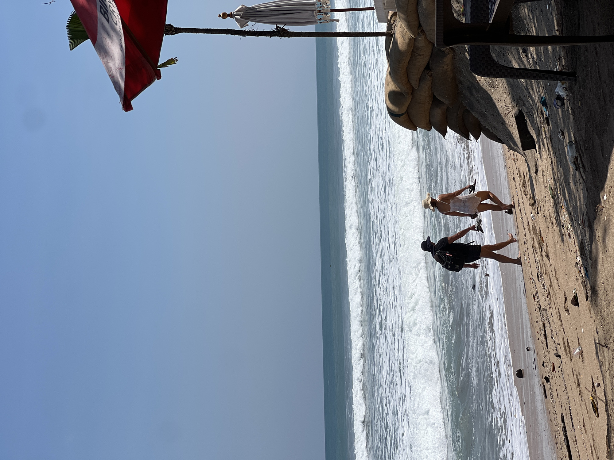 Two individuals walk along the sandy shore of Kuta Beach, Indonesia, with the ocean's waves crashing in the background. The person on the left is wearing a dark t-shirt, shorts, a hat, and a backpack, carrying flip-flops in their hand. The person on the right is wearing a white sundress and a straw hat, also carrying flip-flops. To the right of the scene, sandbags are stacked, and a partially visible red umbrella and a closed white umbrella are erected on poles. Scattered debris, including plastic bottles and driftwood, litters the beach. The sky is clear and blue, and the sun appears to be high in the sky, casting shadows on the sand.