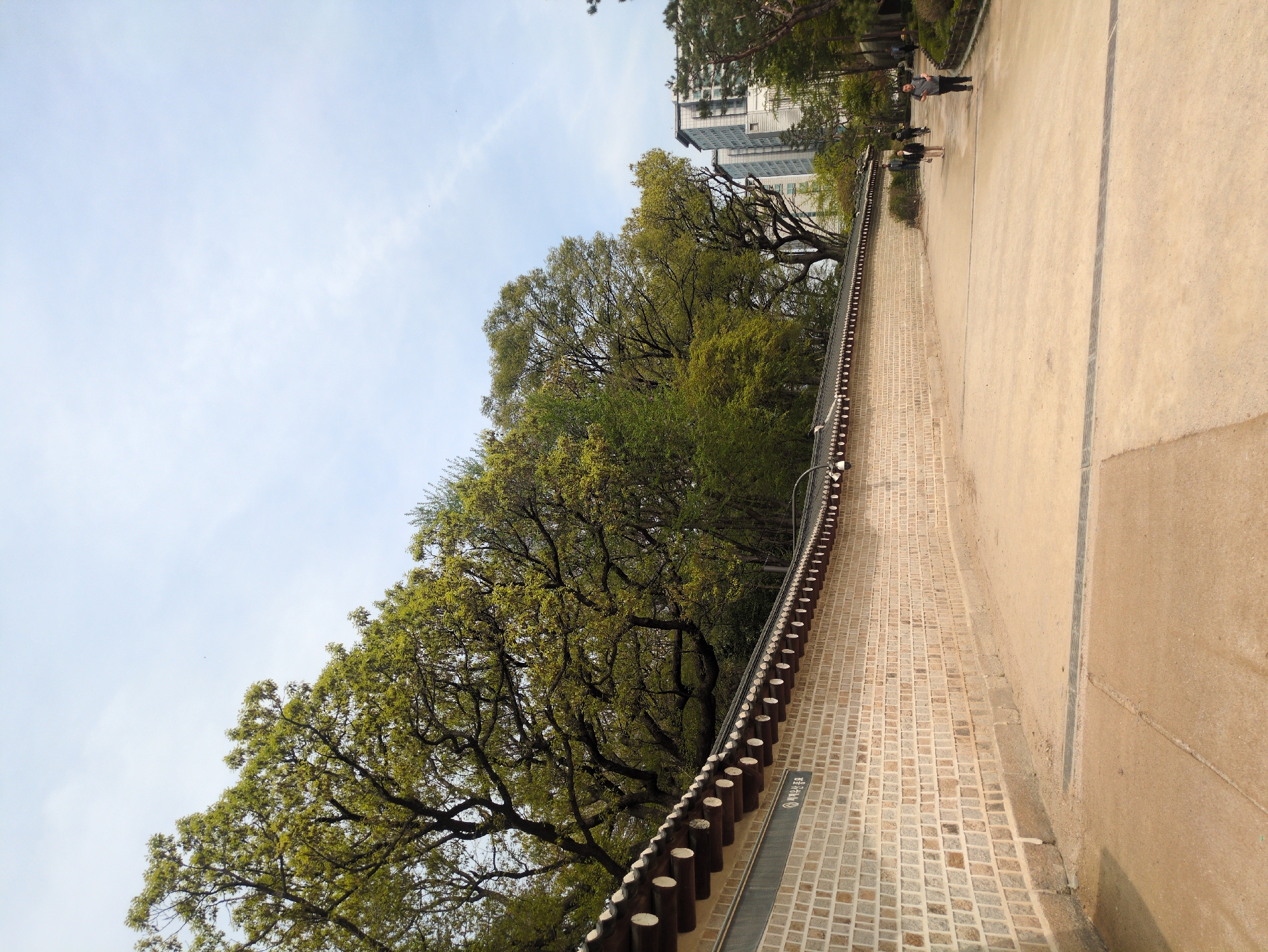 The image depicts a paved pathway alongside a traditional Korean brick wall with a wooden fence on top. Trees with green foliage overhang the wall. In the background, modern apartment buildings are visible. Several people are walking along the pathway. The scene is likely located in a park or historical area within the city of Seoul, South Korea.