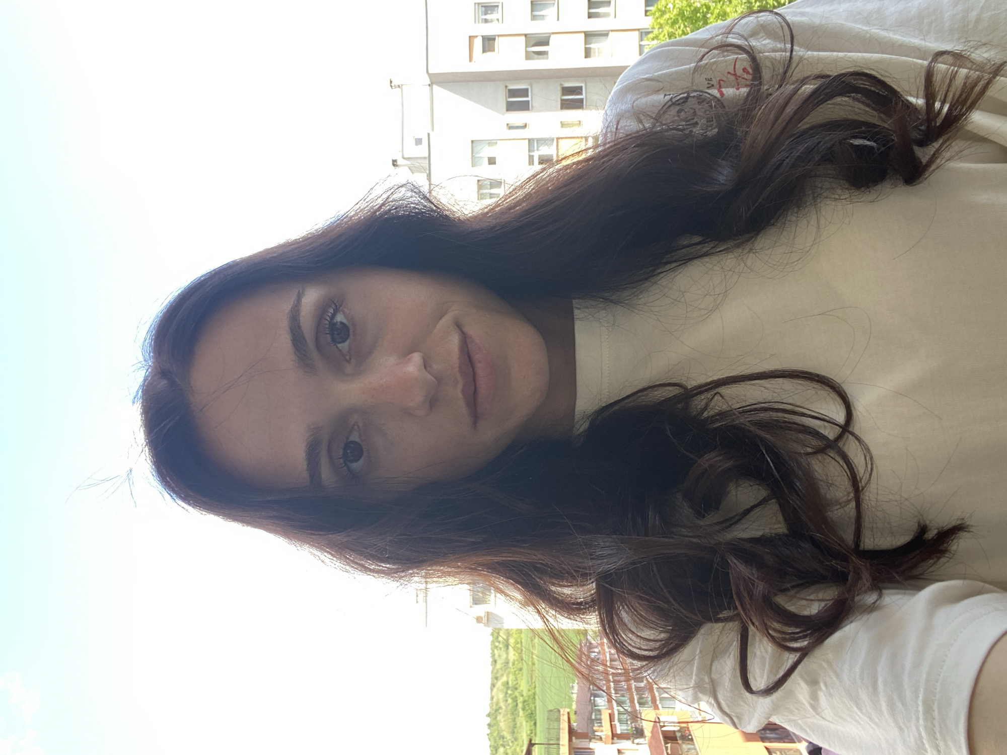 A young woman is posing for a selfie, indoors but with an outside view that suggests a balcony setting. She has long, dark brown hair, fair skin, and is wearing a light-colored t-shirt. Behind her is a building and trees, which appear to be in Blagoevgrad, Bulgaria. The setting appears to be daytime.