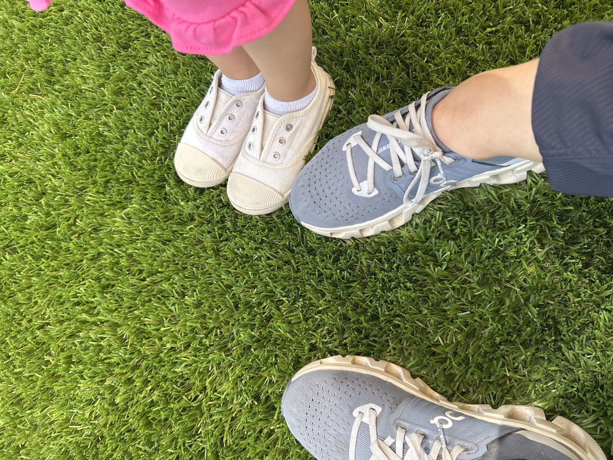 The image shows two pairs of shoes on a green, grassy surface, likely artificial turf. In the upper left corner, a child's feet are visible, wearing white socks and light-colored canvas shoes with no laces. To the right of the child's shoes, an adult is wearing a gray and white running shoe with laces and a glimpse of navy-blue fabric above the ankle. In the bottom center of the frame is the adult's second running shoe. The setting appears to be outdoors. The image was taken in Puebla, Mexico.