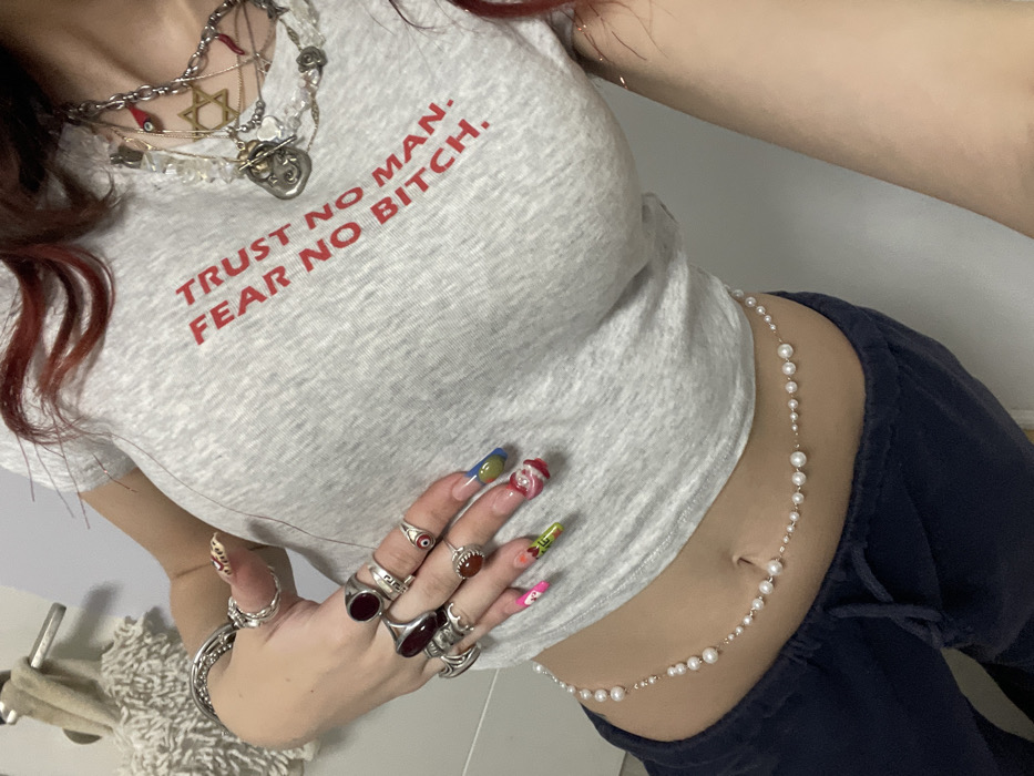 The image displays a close-up, cropped view of an individual's torso, left arm, and hand in an indoor setting. The individual, who has reddish-brown hair visible on the right, wears a light grey, short-sleeved crop top with the red text "TRUST NO MAN. FEAR NO BITCH." printed on the chest. Below the crop top, dark blue sweatpants with a drawstring are visible, extending from the lower abdomen.
The left hand is adorned with multiple rings, including large silver-toned rings set with dark red and orange-brown stones, and one with an evil eye design. The fingernails on the left hand are long and display diverse, colorful artistic designs. Several silver-toned bracelets are stacked on the left wrist. Around the neck, multiple layered necklaces are worn, featuring various charms such as a Star of David, a hamsa-like pendant, and a red horn, alongside a necklace composed of clear or white beads. A delicate waist chain, made of white pearls of varying sizes strung on a thin metallic chain, encircles the individual's upper abdomen, above the waistline of the sweatpants.
The background consists of a plain, light-colored wall or door, with a metallic handle visible in the upper left corner. The specific location within the city of Raanana, Israel, cannot be determined from the visual information presented in the image.