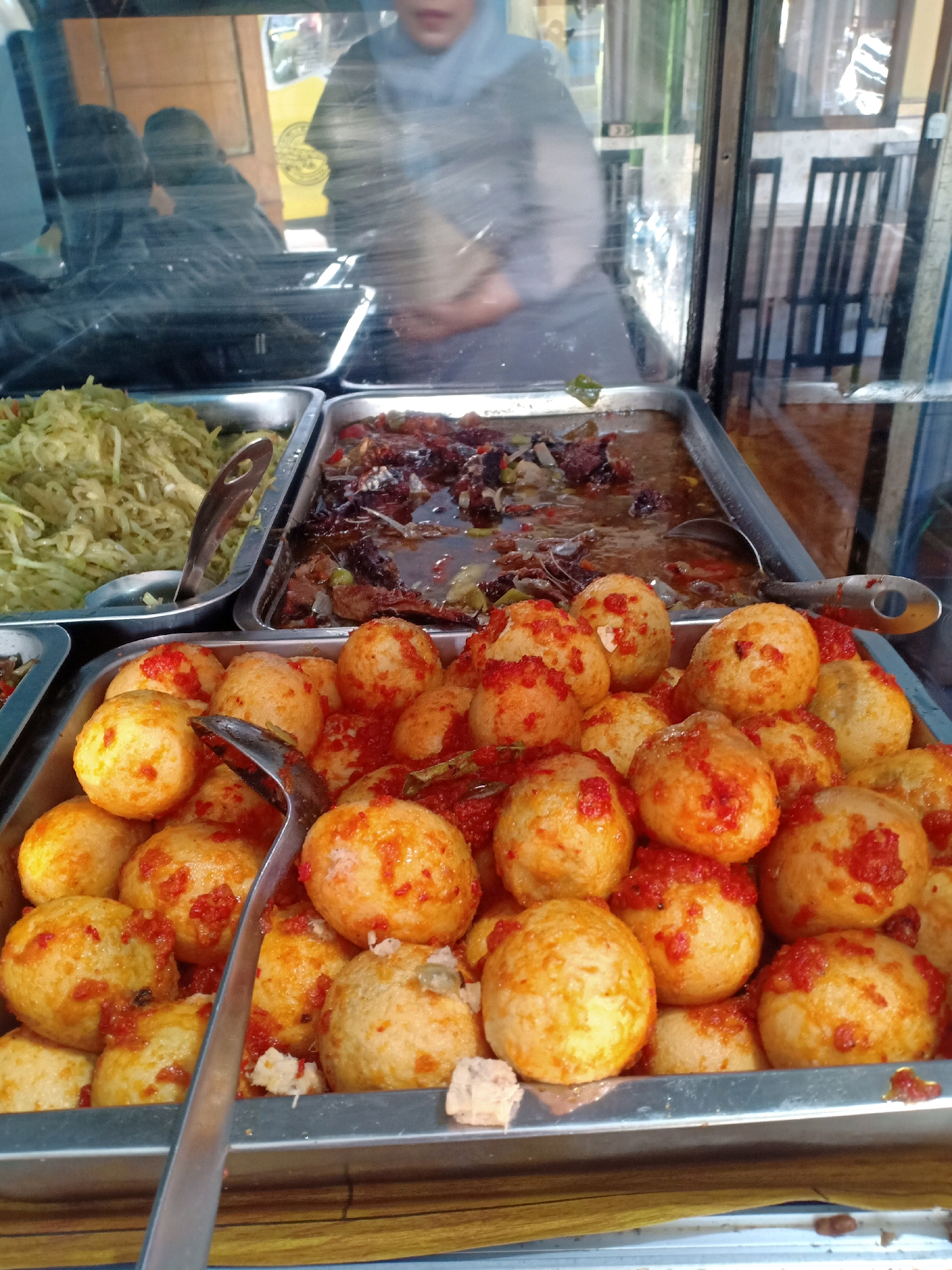 A food stall display is shown, featuring several metal trays of Indonesian cuisine. In the foreground, a tray is filled with numerous small, round, golden-brown fried items, likely quail eggs, coated in a reddish sauce. A silver serving spoon rests diagonally across this tray. To the left, another tray holds a mound of shredded green vegetables, possibly cucumber or papaya salad, with a metal serving spoon. Behind these, a tray contains a dark, saucy dish with pieces of meat and vegetables. A glass partition separates the food from the viewer. Reflections in the glass show two people, one with their back to the camera wearing a light-colored headscarf and grey shirt, and another figure partially visible beside them. In the background, through the glass, a checkered pattern of chairs and tables is discernible, suggesting an indoor or covered seating area. The overall impression is a vibrant, local food vendor.