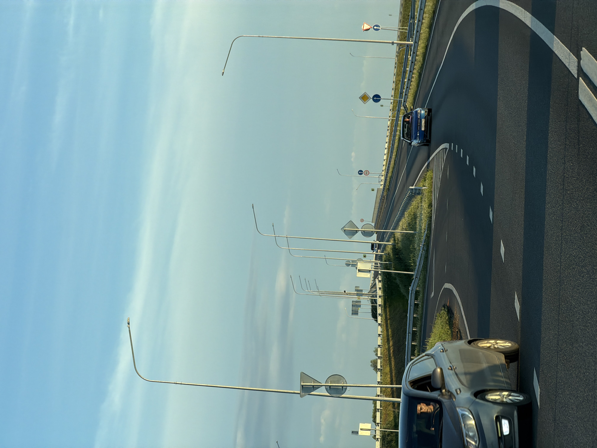 This is a medium-shot, eye-level, vertical photograph taken on a road in Obukhovo, Russia. The sky is a clear, pale blue with some wispy clouds. The road is asphalt and curves to the right. There are several cars on the road. In the foreground, a gray car is in the left lane, and a blue car is in the right lane. The blue car has a license plate that appears to read "Y493MO 190". Both cars have drivers visible. There are streetlights and various road signs lining the road. The environment is rural, with grassy verges and distant trees. The sun appears to be low in the sky, casting long shadows.
