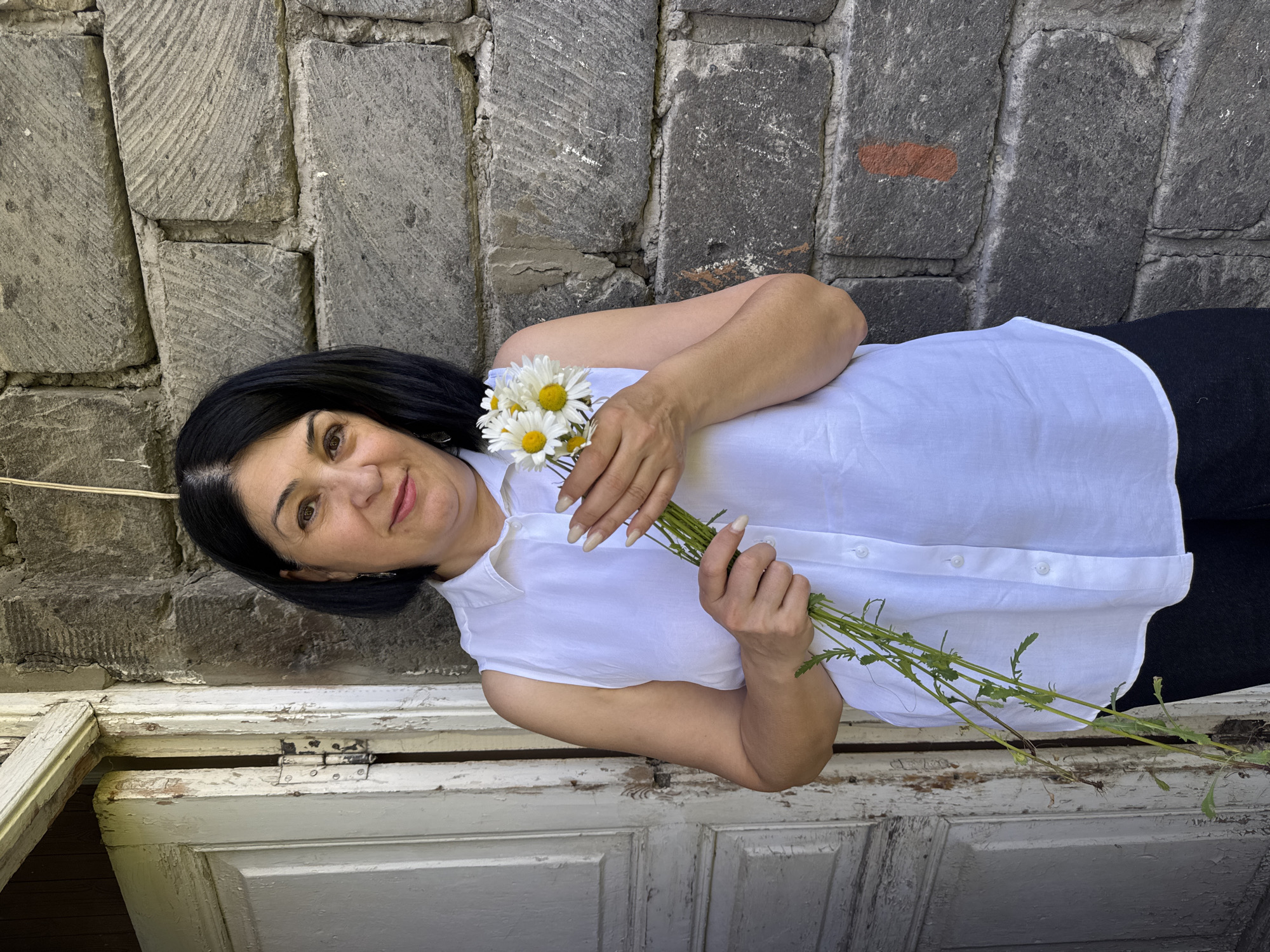 A woman stands in front of a stone wall and a dilapidated wooden door. She has dark hair and is wearing a white sleeveless shirt and black pants. In her hands, she holds a small bouquet of daisies. The stone wall appears to be part of an old building, and the door is weathered and peeling paint. The overall impression is one of a rural or historical setting, possibly in Gyumri, Armenia, given the architecture. The lighting suggests it is daytime.