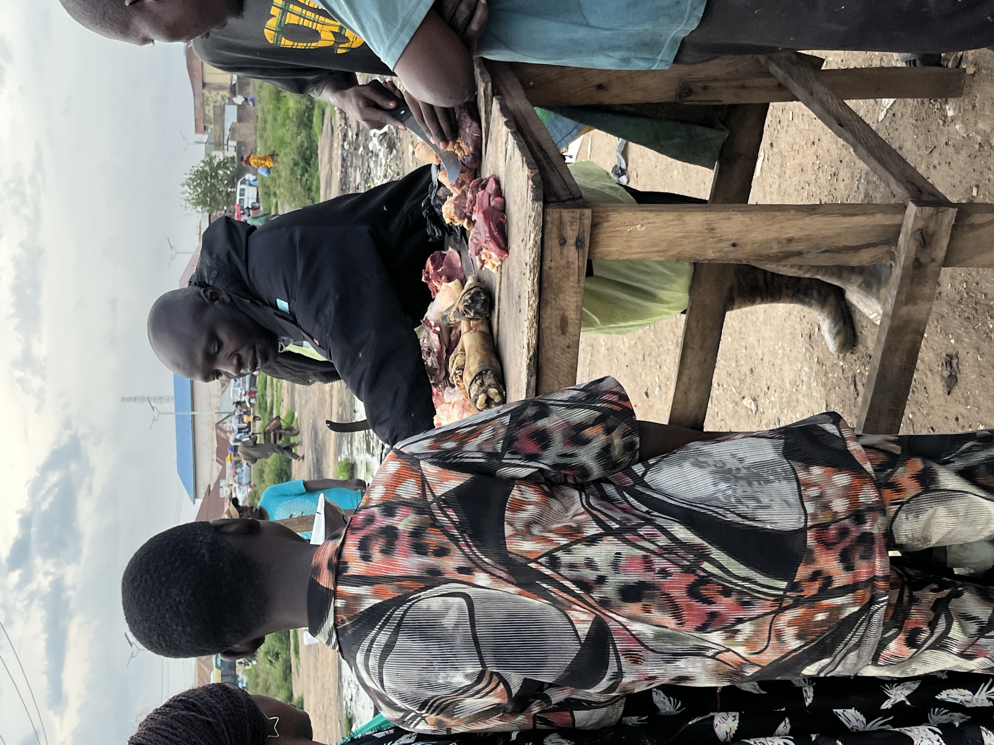 In Bukuru, Nigeria, at an outdoor market, a butcher, wearing a black jacket and patterned trousers, cuts meat on a wooden table. He is surrounded by potential customers. A young person in a vibrantly patterned shirt stands opposite him, while another person, wearing a blue t-shirt, stands to the right, observing the butcher's work. The market appears to be in a state of disarray, with debris scattered on the ground. The sky is overcast, suggesting a cloudy day. The overall scene depicts a typical day at a bustling Nigerian market.