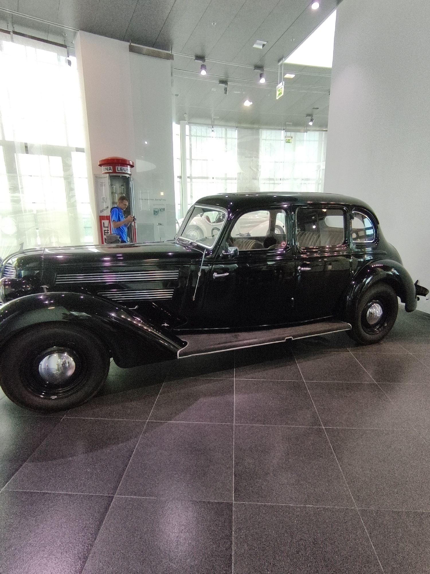 An old black sedan is parked on a tiled floor in a well-lit museum hall. The car has chrome accents and a spare tire mounted on the rear. To the left of the car is a vintage gas pump with red and white branding. A man wearing glasses and a blue shirt is standing near the gas pump, looking at his phone. The environment appears to be an indoor exhibition space with large windows in the background, suggesting it might be part of the Audi museum in Ingolstadt, Germany, given the historical context of the car and brand.