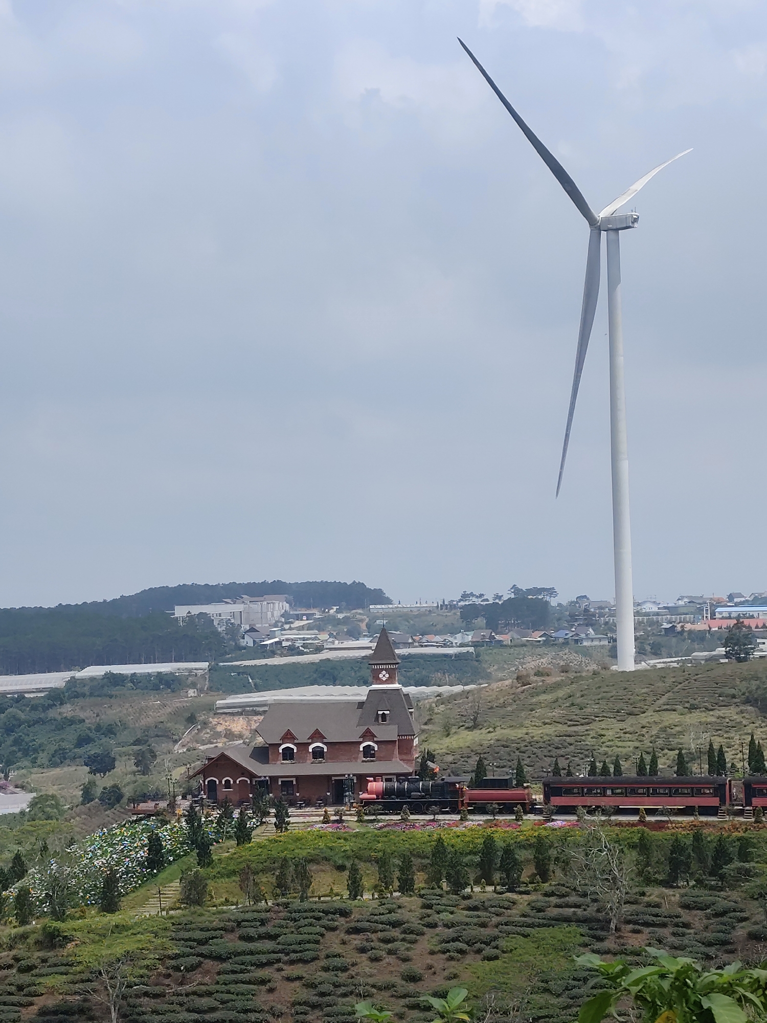 The scene depicts a landscape with a building resembling European architecture, a train, and a wind turbine. The building is a two-story structure with a tower, red brick walls, and arched windows. A black steam locomotive and red train cars are parked along the tracks. A large, white wind turbine dominates the landscape, extending upwards into the cloudy sky. The foreground consists of a lush, green hillside with tea plants and flower bushes. In the background, there are houses and greenhouses in the distance. This is in Trường Thọ, Vietnam.