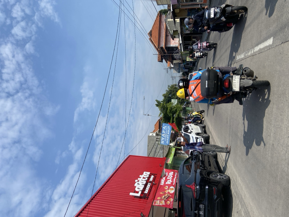 The scene takes place on a street in Kecamatan Tanggulangin, Indonesia, located at latitude -7.5 and longitude 112.702. The street is paved with asphalt and lined with shops and buildings on both sides. The sky is blue with some white clouds. There are several vehicles parked along the street, including a black SUV, a white sedan, and a motorcycle. There are also several people walking and riding motorcycles on the street. A man in a white shirt and blue jeans is walking on the street. He is waving his left hand in the air. There is a sign that reads "SYAFA' AT. WORN A. JAKET KULIT." Two people are riding a motorcycle down the street. The person in the back is wearing a blue and yellow hard hat. The motorcycle has a black and silver top case. Several other motorcycles and people are visible in the distance. The scene is bright and sunny with a few light-colored clouds in the sky.