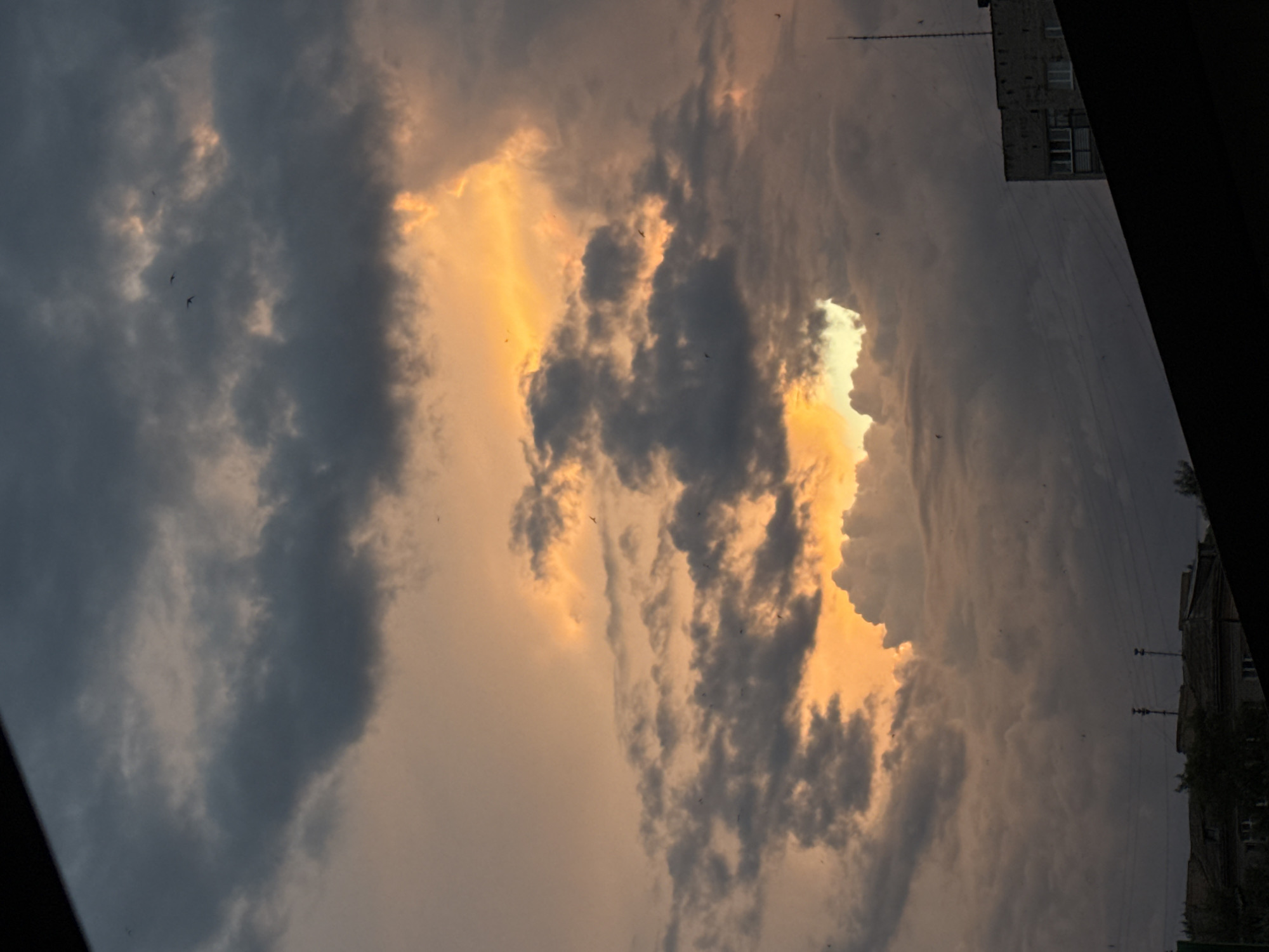 A sunset casts an orange glow through breaks in dark, dramatic clouds, creating a striking contrast in the sky above Luhansk, Ukraine. Below the sky, the silhouettes of urban rooftops, including a multi-story apartment building with visible windows and an antenna, are visible against the darkening horizon. A dark foreground element, likely a roofline or other structure, partially obscures the lower part of the frame. Small birds are scattered throughout the sky.