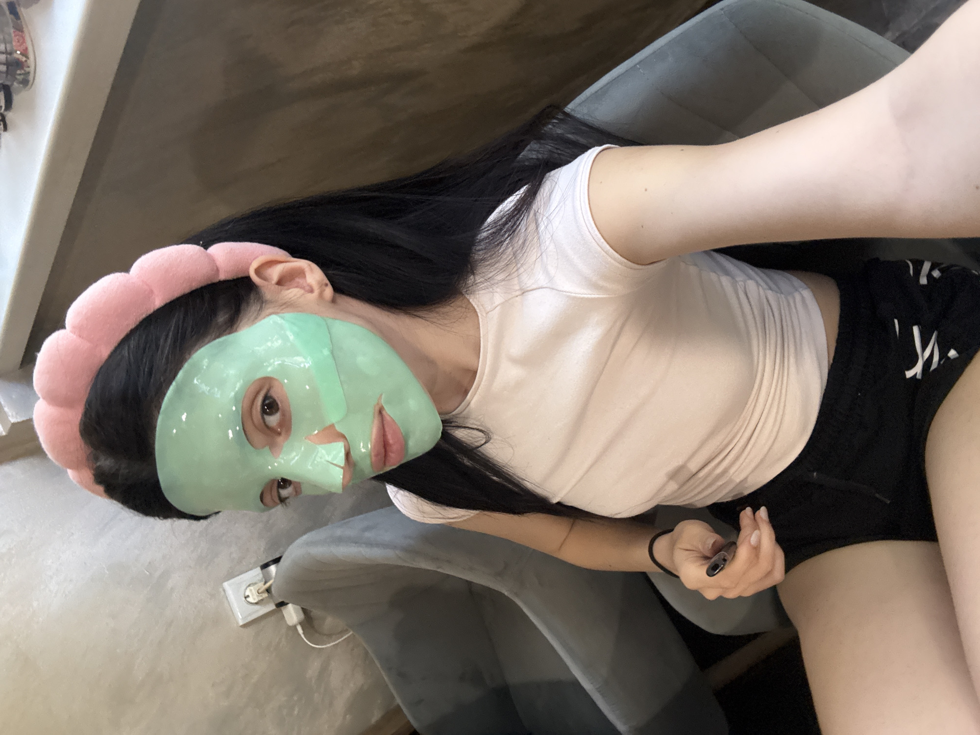 A woman is sitting in a gray chair with a green facial mask and a pink headband. She has long, black hair, and is wearing a white t-shirt and black shorts. She is holding a small silver device in her right hand. A wall outlet and charging cables are visible near the chair. The location cannot be determined from the image.