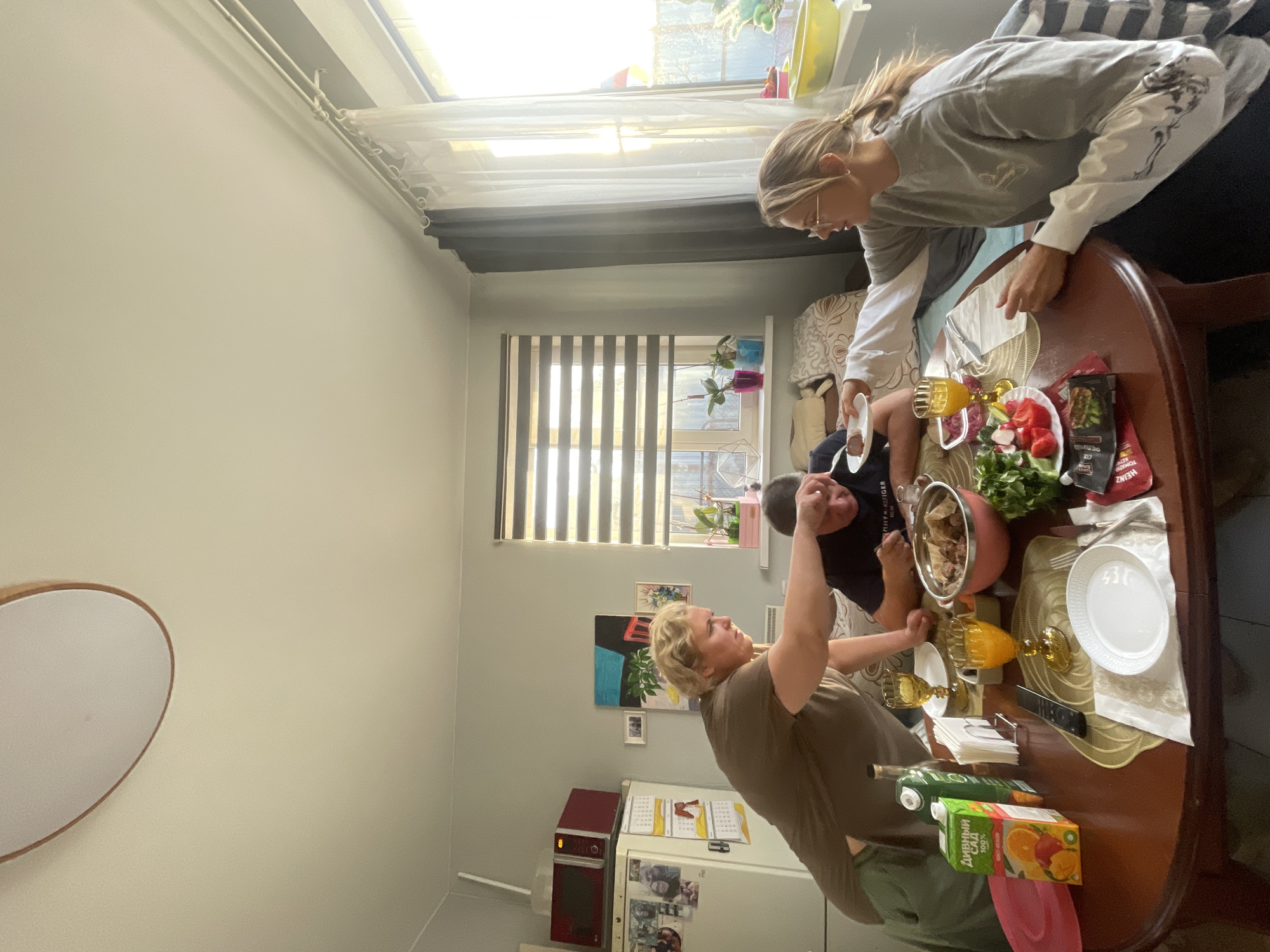 A family is gathered around a brown, oval-shaped wooden table, indoors, likely in a home in Sestroretsk, Russia. Three people are visible: a woman with short blonde hair wearing a brown t-shirt, a younger person with dark hair in a dark t-shirt, and a young woman with blonde hair in a light gray sweater.
The woman with blonde hair is bent over the table, holding a white plate. The person with dark hair appears to be reaching towards a pink pot at the center of the table. The woman with short blonde hair is facing the table, seemingly serving the food.
The table is covered with a beige cloth. Dishes, cutlery, glasses of orange juice, a bowl of salad, ketchup, and orange juice cartons are present on the table.
Behind the table is a window with white curtains and vertical blinds. A refrigerator with a microwave on top, and photos attached, is visible in the background. A piece of artwork is hung on the wall, and a round mirror is hanging on the wall to the left.