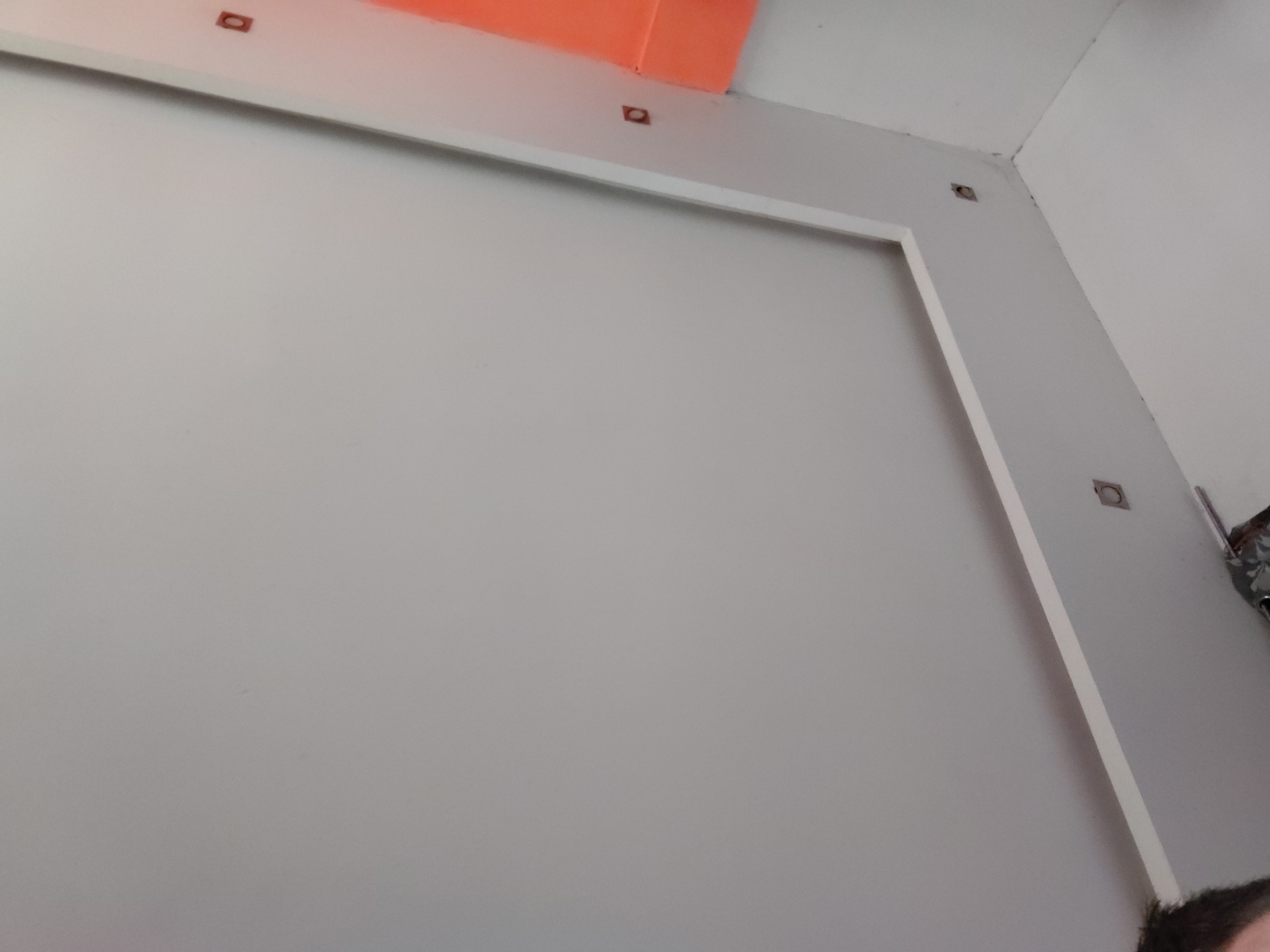 The image presents an upward-angled view of an interior space, likely a room under construction or renovation in Santa Ana, Venezuela. The central subject is a light grey wall adorned with a prominent white rectangular trim or frame. Above this wall, a narrow white section, possibly part of the upper wall or ceiling, features several small, dark, rectangular inserts or holes, which may serve as mounting points for fixtures. In the upper left corner, a striking bright orange object, possibly a pipe, conduit, or insulation, stands out against the muted palette. The right side of the frame shows an interior corner where two white surfaces meet, with some dark marks or discoloration along the seam. A small section of a person's head with dark hair is visible in the bottom right corner, indicating human presence. The lighting suggests an indoor environment, likely illuminated by artificial sources. No text is visible in the image.