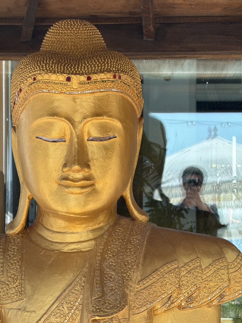 The image displays a close-up of a golden Buddha statue, depicted from the chest up with its eyes peacefully closed in meditation. The statue features an elaborate headpiece (ushnisha) with a decorated band set with small red and clear jewel-like embellishments. Its robe also boasts a finely embossed pattern, highlighted by tiny clear dots that resemble jewels or mirrors, particularly noticeable on the shoulders and chest.
The Buddha is positioned indoors, seemingly behind a large pane of glass, as indicated by a prominent reflection. In this reflection, a person, likely the photographer, is visible wearing sunglasses and holding a camera or phone to capture the image. The outdoor environment reflected in the glass includes a bright blue sky and a distinct large white umbrella or pagoda-like structure to the right of the reflected person. Above the glass, dark wooden beams suggest a ceiling or overhang.
The strong reflections and bright sky imply the photograph was taken on a sunny day. The scene creates a juxtaposition between the serene, traditional religious art of the Buddha and the modern presence of the photographer and the contemporary outdoor setting. Given the location context of Sant Antoni de Portmany, Spain, this statue likely serves as a decorative element in a shop, restaurant, private residence, or garden within the town. No visible text is present in the image.