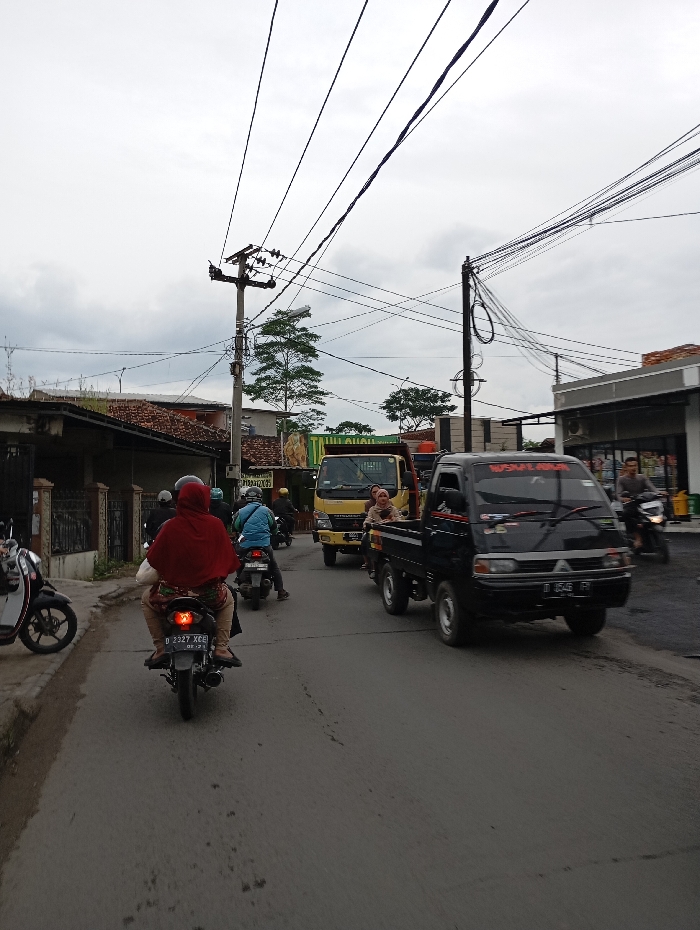 The scene is a narrow, paved street in Kecamatan Kutawaringin, Indonesia. It is daytime, and the sky is overcast. The street is lined with buildings on both sides. Several motorbikes are parked or traveling on the road, one of which is being ridden by a woman wearing a red hijab. The road is relatively empty, with little traffic. There are several power lines overhead. A large truck is parked at the end of the road. A few people are visible, including one woman in a red hijab and another woman standing next to the large truck. Several trees are visible on the left side of the road.