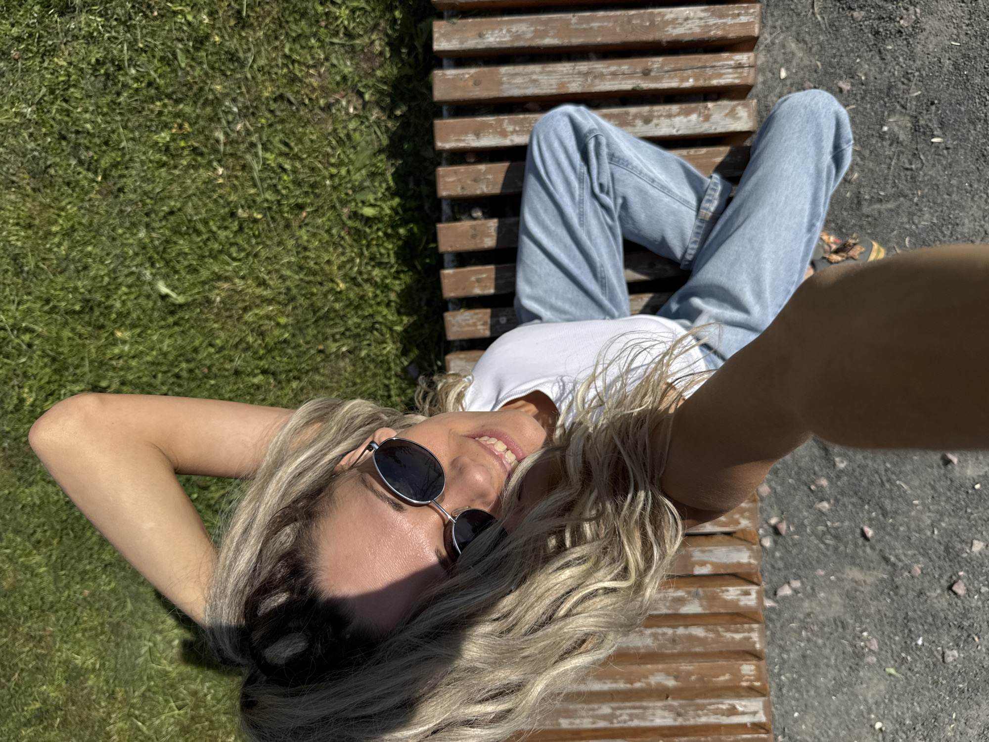 A woman with blonde, wavy hair pulled back in a ponytail, wearing sunglasses, a white t-shirt, and light blue jeans, is sitting on a wooden park bench. She is smiling and holding her arm behind her head, as if posing for a selfie. The bench is situated on a grassy area, with dirt and gravel visible in the foreground. The lighting suggests it is a sunny day. The location is Rusanovo, Russia.