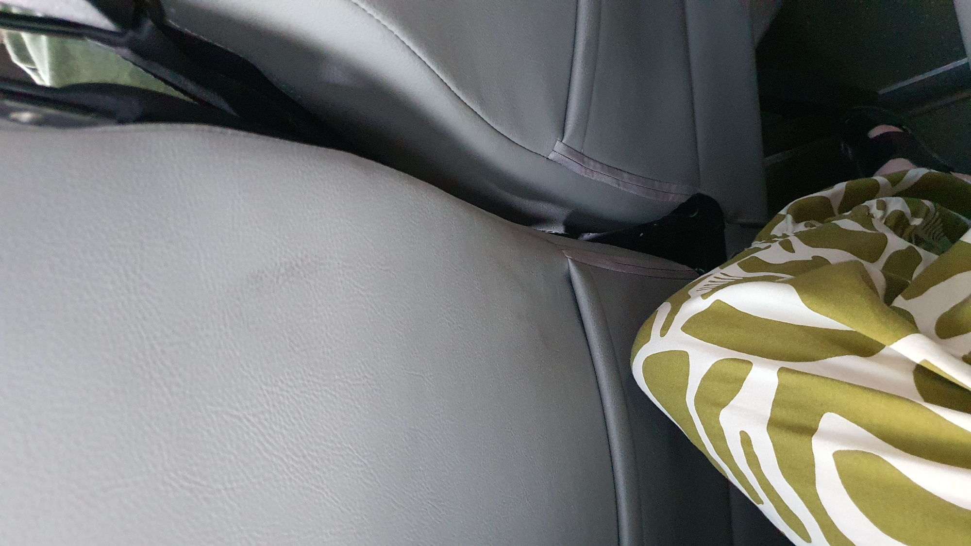The media file shows a close-up view of what appears to be the interior of a vehicle, possibly a bus or a van, given the arrangement of the seats. The dominant colors are various shades of gray from the seats, and a vibrant olive green and white patterned fabric.
In the foreground and occupying the lower right portion of the frame is a crumpled piece of fabric with a bold, abstract pattern resembling stylized animal print or organic shapes. The fabric is a bright olive green with irregular white shapes. It's positioned on what seems to be a seat.
Behind the patterned fabric, and extending across the majority of the image, are gray seats. The seats are made of a material that looks like faux leather or vinyl, with visible stitching lines indicating their construction. The perspective suggests the viewer is seated or looking from a similar vantage point within the vehicle.
In the upper right corner, a dark-colored shoe or sandal is partially visible, indicating a person's presence. A person's legs are also visible, clad in what appears to be dark-colored leggings or trousers, partially covered by the patterned fabric.
The lighting suggests it is daytime, but the interior setting makes it difficult to determine the exact time of day or weather conditions. There are no discernible location cues beyond the vehicle interior, and no visible text. The overall impression is a candid snapshot taken from within a moving or stationary vehicle, focusing on the textures and colors of the seats and a piece of fabric. The scene is neutral in terms of expressed emotion.