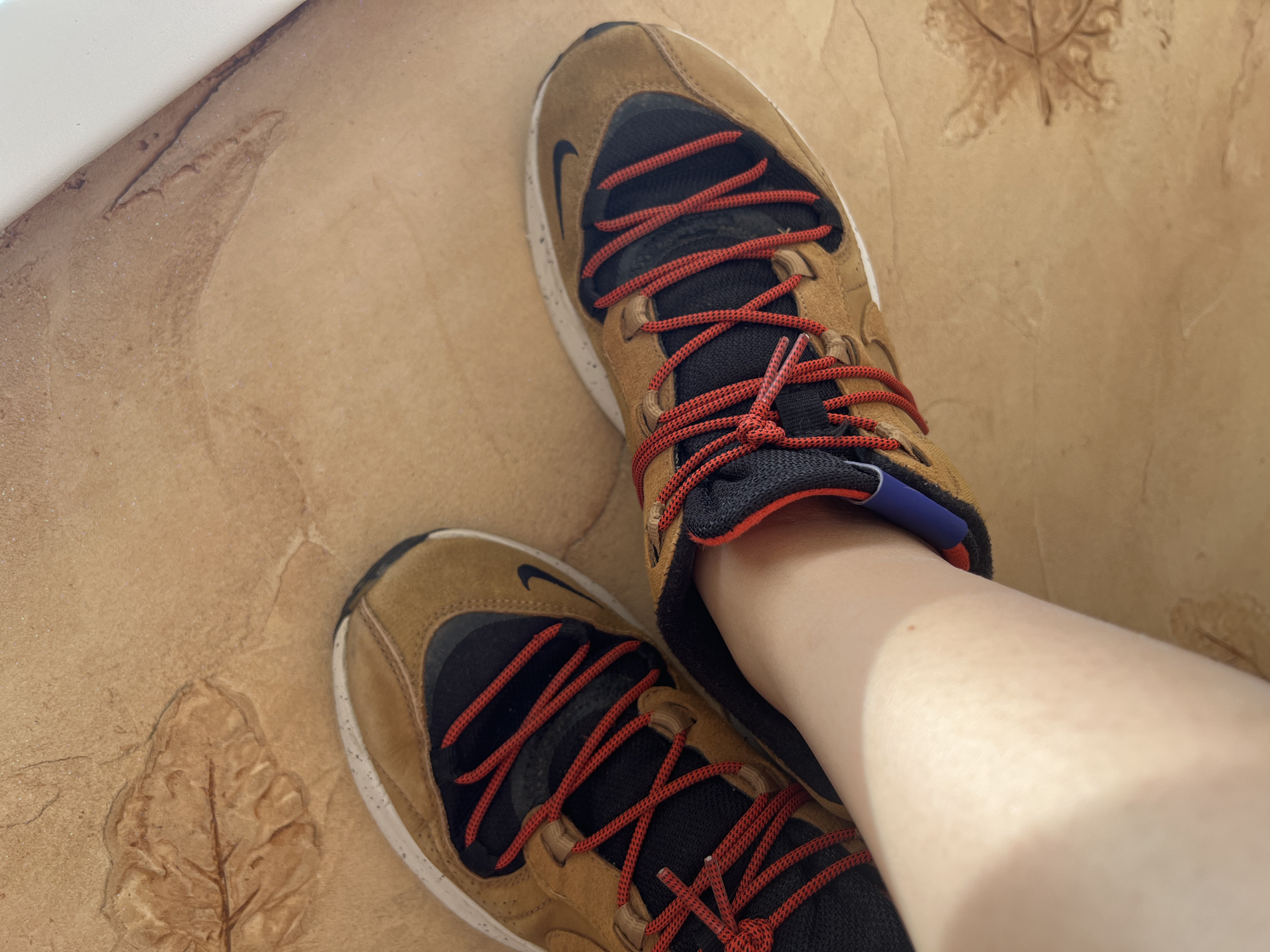 The image shows a person's feet wearing brown and black Nike sneakers with red laces. The sneakers are on a light brown tiled floor with embossed leaf patterns. The scene is lit by natural light, casting shadows. The background is blurred, but appears to be an indoor setting. There is no discernible information in the image to suggest a specific location in Tolyatti, Russia.