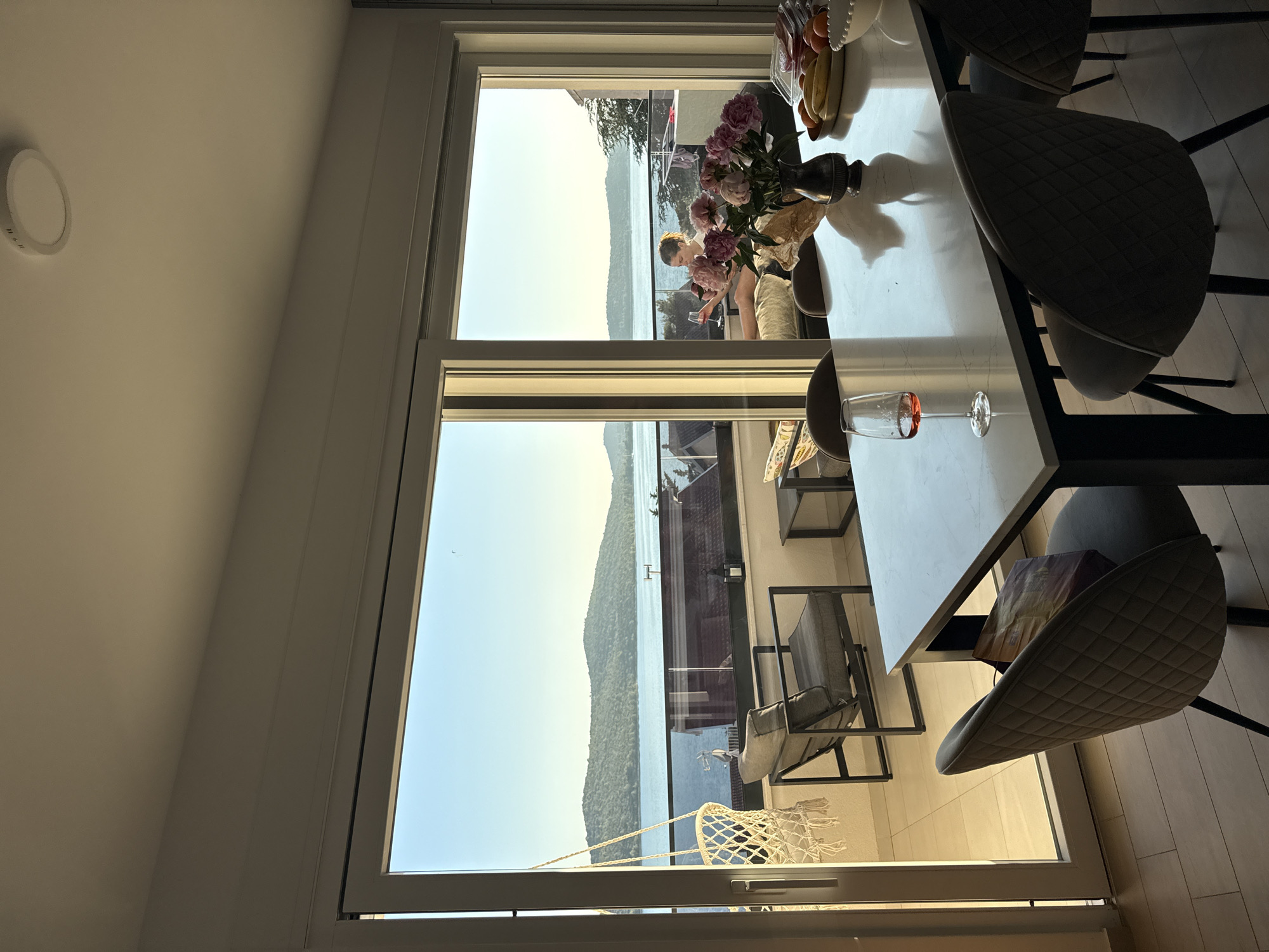 This image depicts an indoor dining area with a large sliding glass door opening to a balcony with a view of the sea and a hilly, tree-covered coastline, likely in or near the city of Šibenik, Croatia. In the foreground, a white marble-topped dining table is set with four upholstered chairs. A glass of rosé wine, a vase of pink peonies, and a fruit bowl containing bananas and oranges are on the table. Through the glass door, a balcony is visible with two chairs, a small table, and a hanging macrame swing chair. A woman is seated on the balcony, holding a glass of wine. The sun is low in the sky, casting a warm glow on the scene.