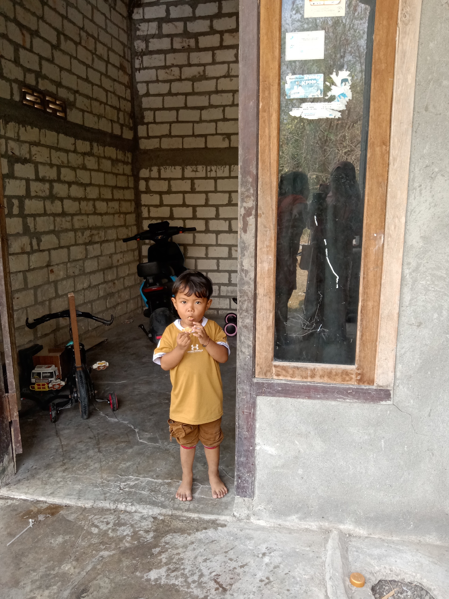 A young boy stands in the doorway of a building, eating something. He is wearing a yellow t-shirt and brown shorts. To his left, a tricycle and a motorcycle are parked. The building appears to be made of concrete blocks, with a wooden-framed window to the boy's right. The ground is concrete and appears to be outdoors. The location is the city of Jumput, Indonesia.