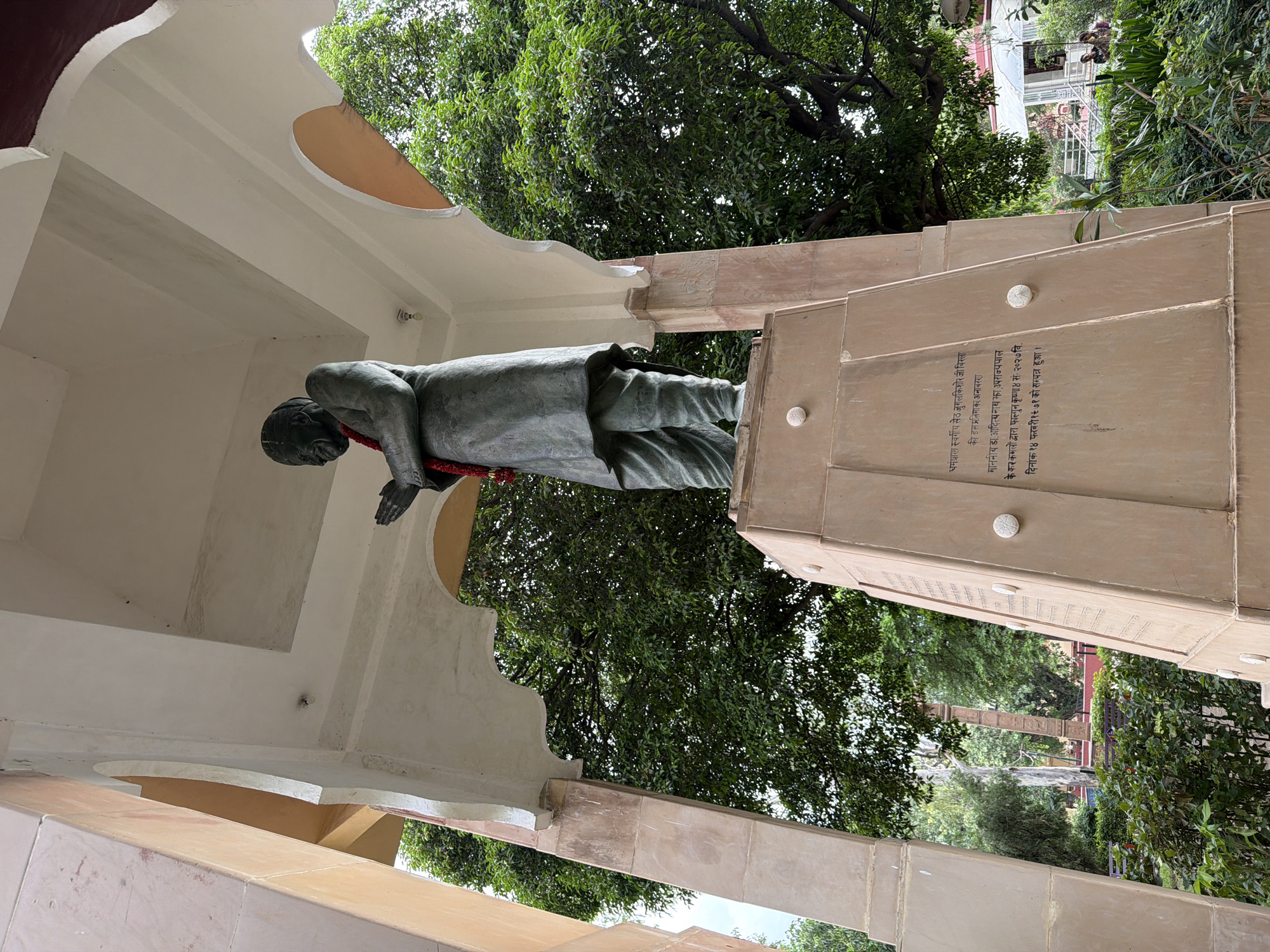 The image depicts a statue of a man in prayer, situated outdoors in what appears to be a park or garden. The statue is made of dark material and is adorned with a red garland. It stands on a tall, tiered pedestal made of light-colored stone, inscribed with text. The surrounding environment includes lush green trees and foliage, with a building visible in the background. The overall scene suggests a memorial or a place of cultural significance. The architectural elements of the structure housing the statue, with its archways and pillars, indicate a deliberate design for public display. The lighting suggests it is daytime, with overcast skies or diffused sunlight contributing to a soft illumination of the scene.