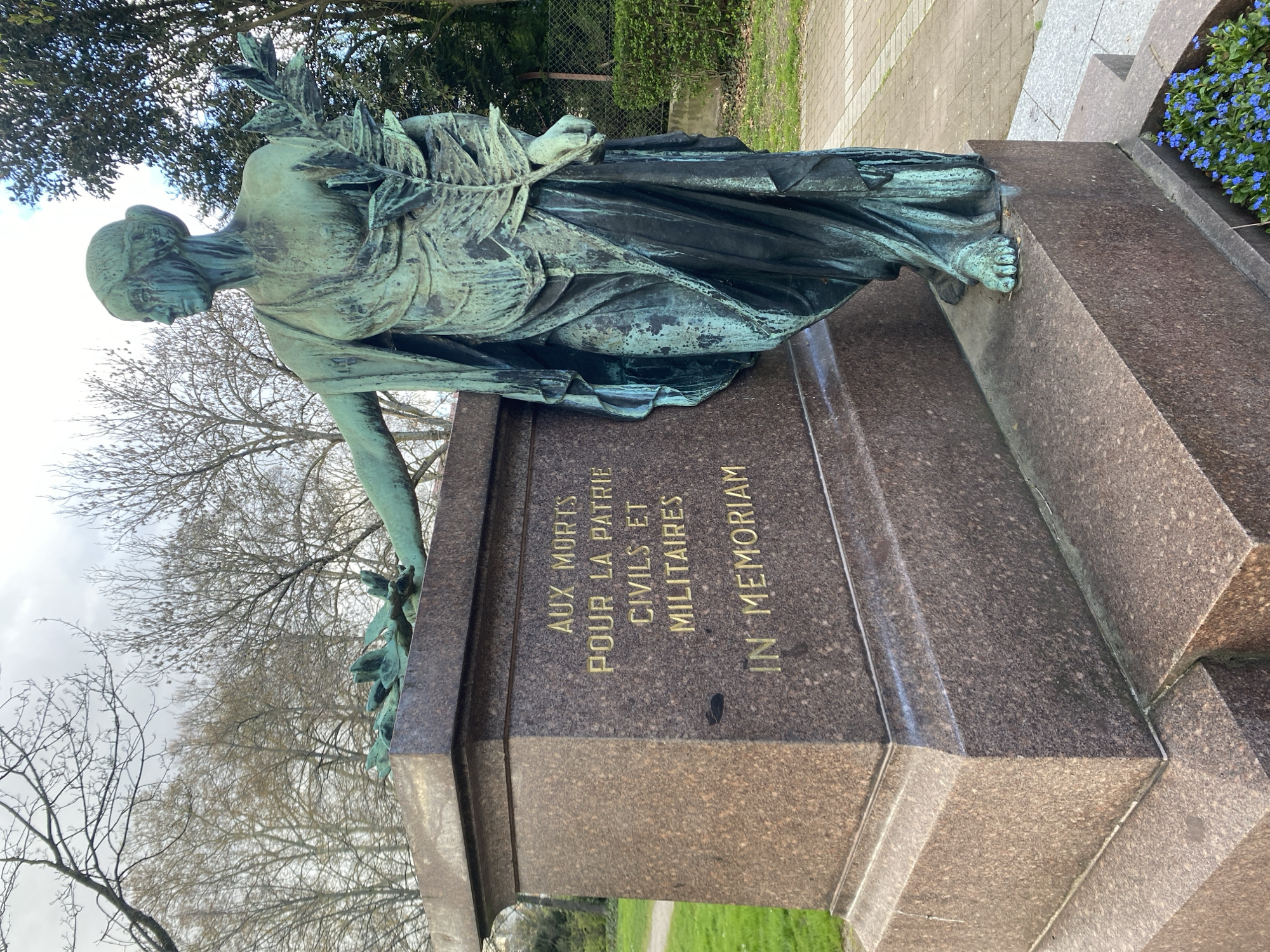 A bronze statue of a draped woman is mounted atop a granite monument. The statue is weathered, with verdigris coloring. The woman has a cloth or cap covering her head, and she holds a branch or frond. The monument is inscribed with the words "AUX MORTS POUR LA PATRIE CIVILS ET MILITAIRES IN MEMORIAM" in gold lettering. The monument is located in a park or cemetery. Trees are visible in the background. The photograph was taken at eye level, slightly to the side of the statue. The location is the city of Schiltigheim, France.