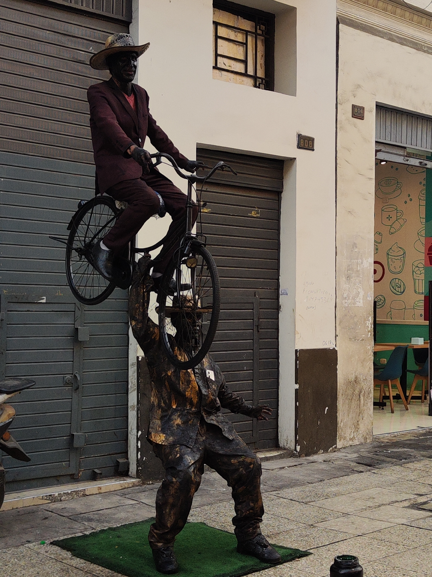 In Lima, Peru, two street performers pose as statues on a city sidewalk. One figure, painted in a dark brown and black, stands on a small patch of green artificial turf, supporting a second performer painted in a similar dark hue, who sits atop a bicycle. The upper performer is clad in a brown suit, a brown hat, and dark gloves, while the supporting performer wears a textured suit of the same color. They are positioned in front of a building with roll-up garage-style doors on the left and the number "808" visible above one doorway. A cafe-style establishment is visible on the right with its interior containing tables and chairs. A jar for tips sits on the sidewalk nearby.