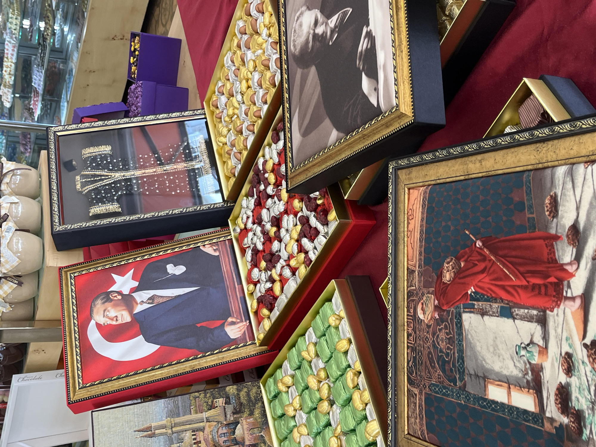 The image shows a display of gift boxes in a store, likely in Alanya, Turkey. The boxes are decorated with images related to Turkish history and culture. Some boxes contain chocolates wrapped in gold, red, green, and silver foil. Several boxes have framed pictures on their lids: one features Mustafa Kemal Atatürk with the Turkish flag, another a man in traditional Islamic dress, a third displays a formal coat, and the fourth a black and white portrait of a man. Purple gift boxes are stacked in the background. The overall scene is a retail setting.