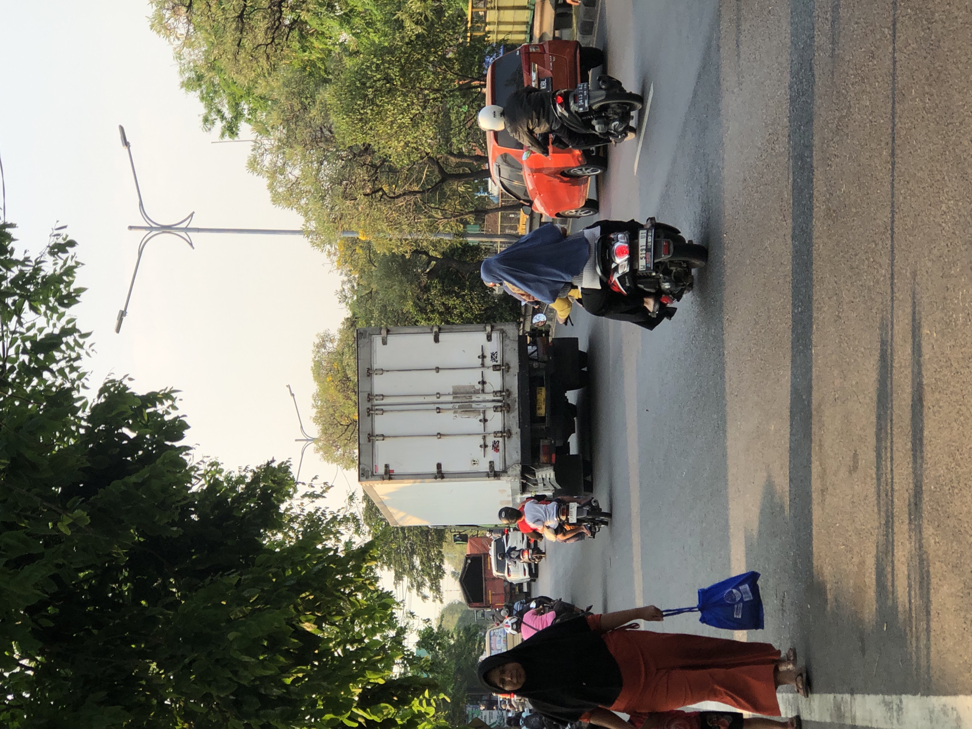 The image depicts a street scene in Kludan, Indonesia, likely during the late afternoon or early evening due to the long shadows cast on the asphalt. The setting is a busy road with several vehicles, including a large white truck, a red car, and numerous motorcycles. Green trees line the road, suggesting a location with ample vegetation.
Several people are visible. A woman in the foreground, wearing an orange skirt and a black headscarf, is holding a blue bag. She is walking on the side of the road. Further down the road, a person on a motorcycle is wearing a blue headscarf. Another motorcyclist in a white helmet and black jacket is seen on a red car's left side. Other individuals are present on motorcycles throughout the scene, indicating ongoing traffic.
The activity appears to be the normal flow of traffic on a public road. The lighting suggests the sun is low in the sky, creating a warm, diffused light. The presence of vehicles and people suggests an urban or semi-urban environment. No specific event is clearly indicated, but the scene captures a moment of daily life on a Indonesian street. Some text is visible on the license plates of the vehicles, but it is not clearly legible.