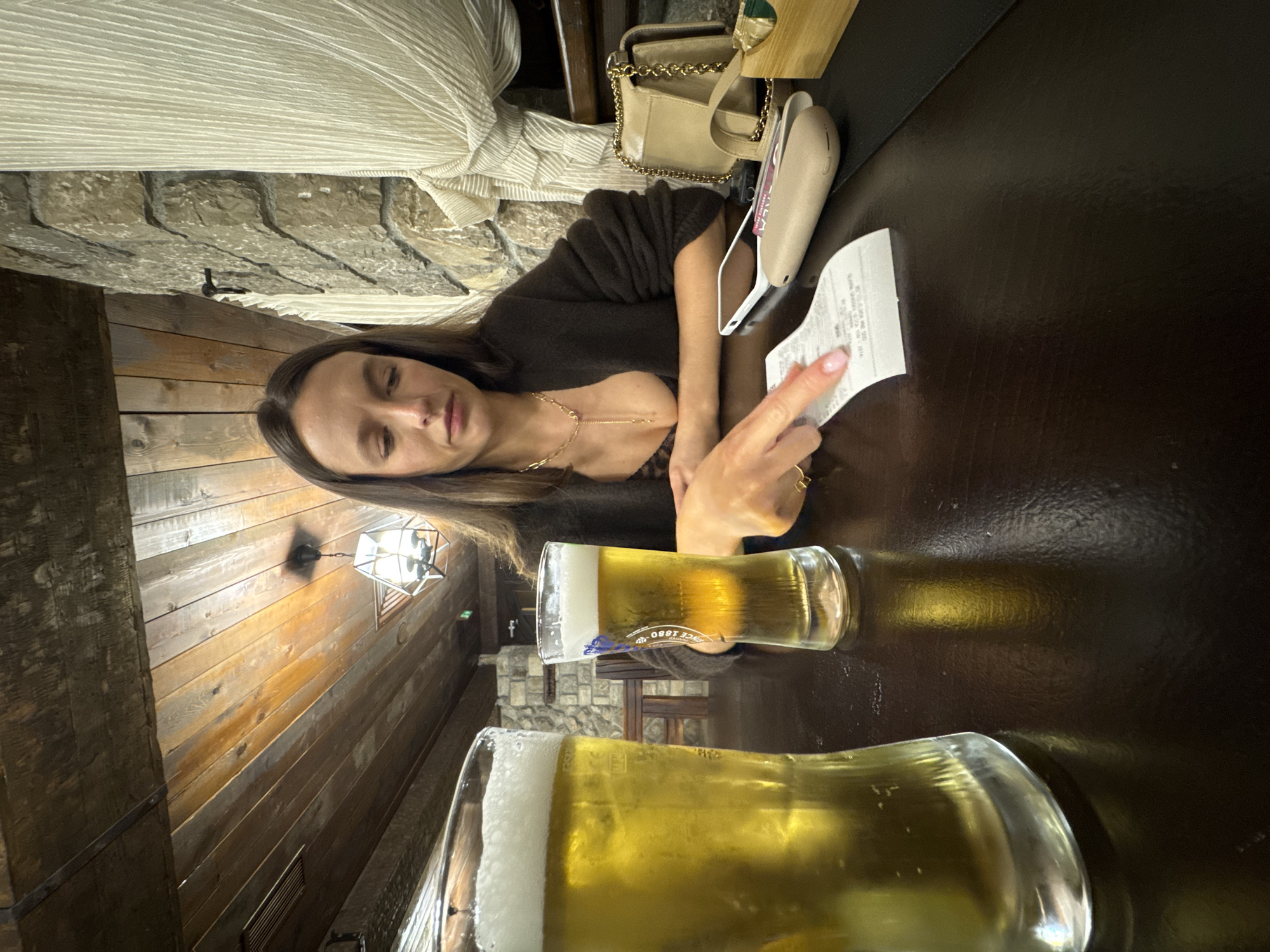 An indoor scene in Mojanovići, Montenegro, depicts a young woman seated at a dark wooden table, holding a receipt in her right hand. The image is oriented vertically, showing the woman from the chest up, with her head resting slightly, eyes closed or looking down. She has long, dark hair and is wearing a dark, possibly knit, long-sleeved top and a delicate gold chain necklace. Her expression is neutral, hinting at contemplation or weariness.
On the table in front of her are two glasses of light-colored beer with foamy heads and condensation, suggesting they are cold. One glass bears partially visible text, including "SINCE 1880". Next to her left arm, a light-colored handbag with a gold chain strap and a beige electronic device (possibly a portable charger or payment terminal) are visible. The receipt she holds is white and appears to contain printed text, though the details are largely illegible.
The background features a rustic interior design. To the left, a textured stone wall is partially visible, topped by light-colored pleated fabric, possibly a curtain or part of the decor. To the right, warm-toned wooden paneling with visible grain covers the wall, and a square, open-caged light fixture hangs from it, casting a soft glow. The overall lighting suggests an evening ambiance. The scene captures a quiet moment, possibly at a restaurant or bar.
