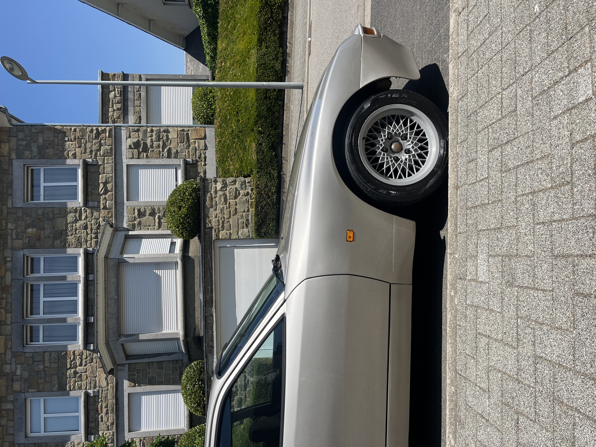 The image shows a beige car parked on a street in front of a stone-faced building. The car is seen from the side, with the front fender and wheel clearly visible. The building features multiple windows with white shutters and a bay window. There is a neatly trimmed hedge in front of the building. A streetlamp stands on the right side of the image. The street is paved, and a cobblestone sidewalk runs along the left side. The sky is clear and blue. The location is identified as Saint-Vith, Belgium.
