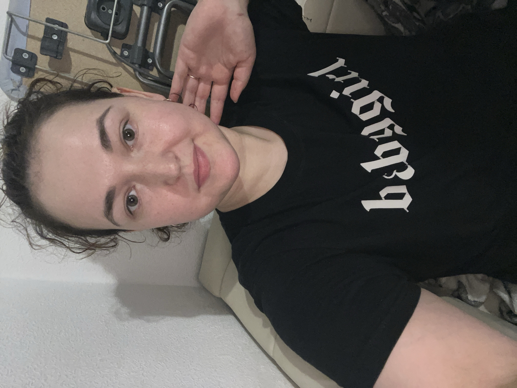 A young woman is shown from the chest up, facing the camera. She has curly brown hair and is wearing a black t-shirt with white gothic lettering that reads "babygirl". Her right hand is raised to her ear, and she is wearing two rings on her fingers and a hoop earring in her visible ear. She appears to be indoors, possibly in a living room, with a neutral-colored sofa or bed behind her. The background also shows a portion of what appears to be a wheeled luggage cart or similar equipment. There is no discernible indication of the city of Chelyabinsk, Russia, or any specific urban location within the image.