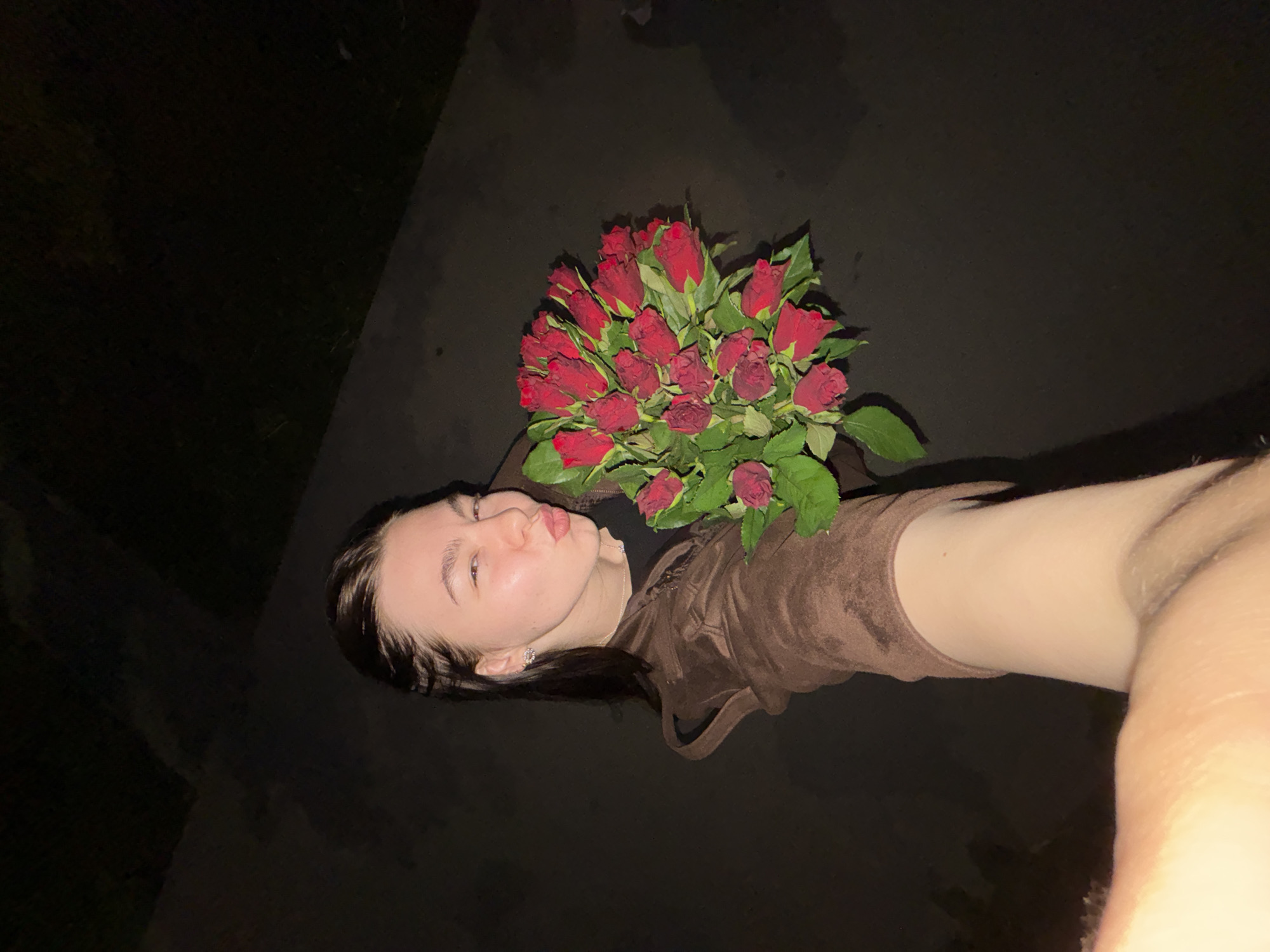 A person is holding a bouquet of red roses. The person is fair-skinned, wearing a brown top, and appears to be taking a selfie. The background is dark, possibly at night, with what appears to be a sidewalk or paved area. There is not enough information to discern the location within Perm, Russia.