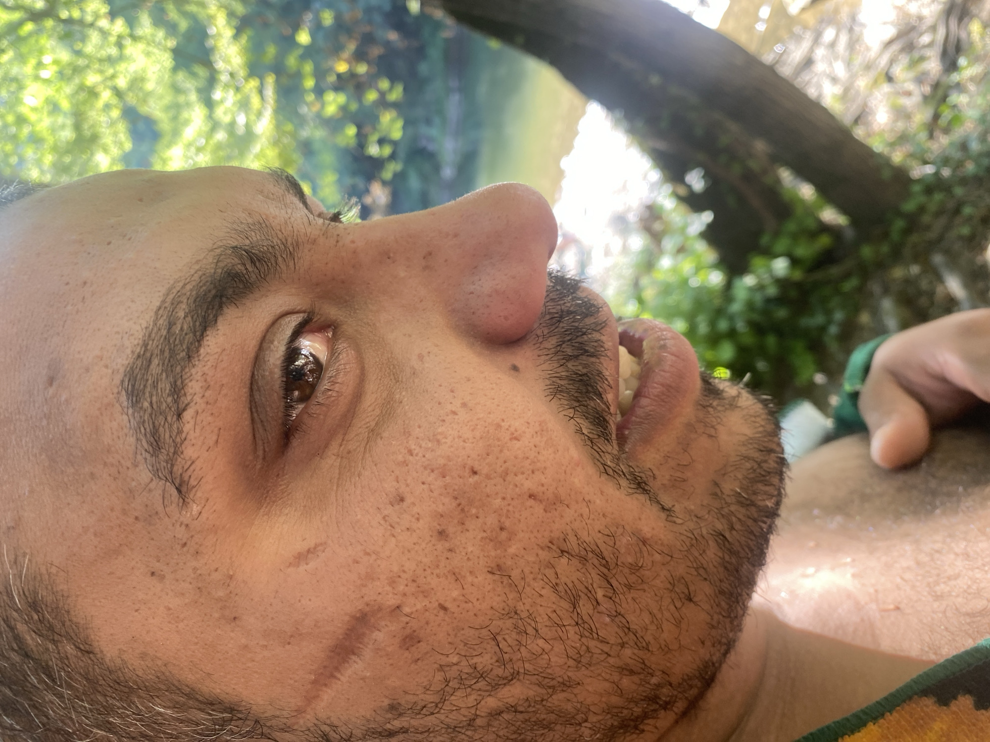The image is a close-up, side profile of a man's face outdoors, with a blurred natural background. The man has brown eyes, a prominent nose, a mustache, and some stubble. There are visible scars and pores on his face. He is wearing a multi-colored garment around his neck. The background shows a body of water and lush green foliage, suggesting a natural setting likely near Debnevo, Bulgaria, a village known for its scenic beauty and proximity to the Yantra River. The lighting indicates it is daytime, with sunlight filtering through the leaves. The scene captures a moment of rest or observation in a natural environment.