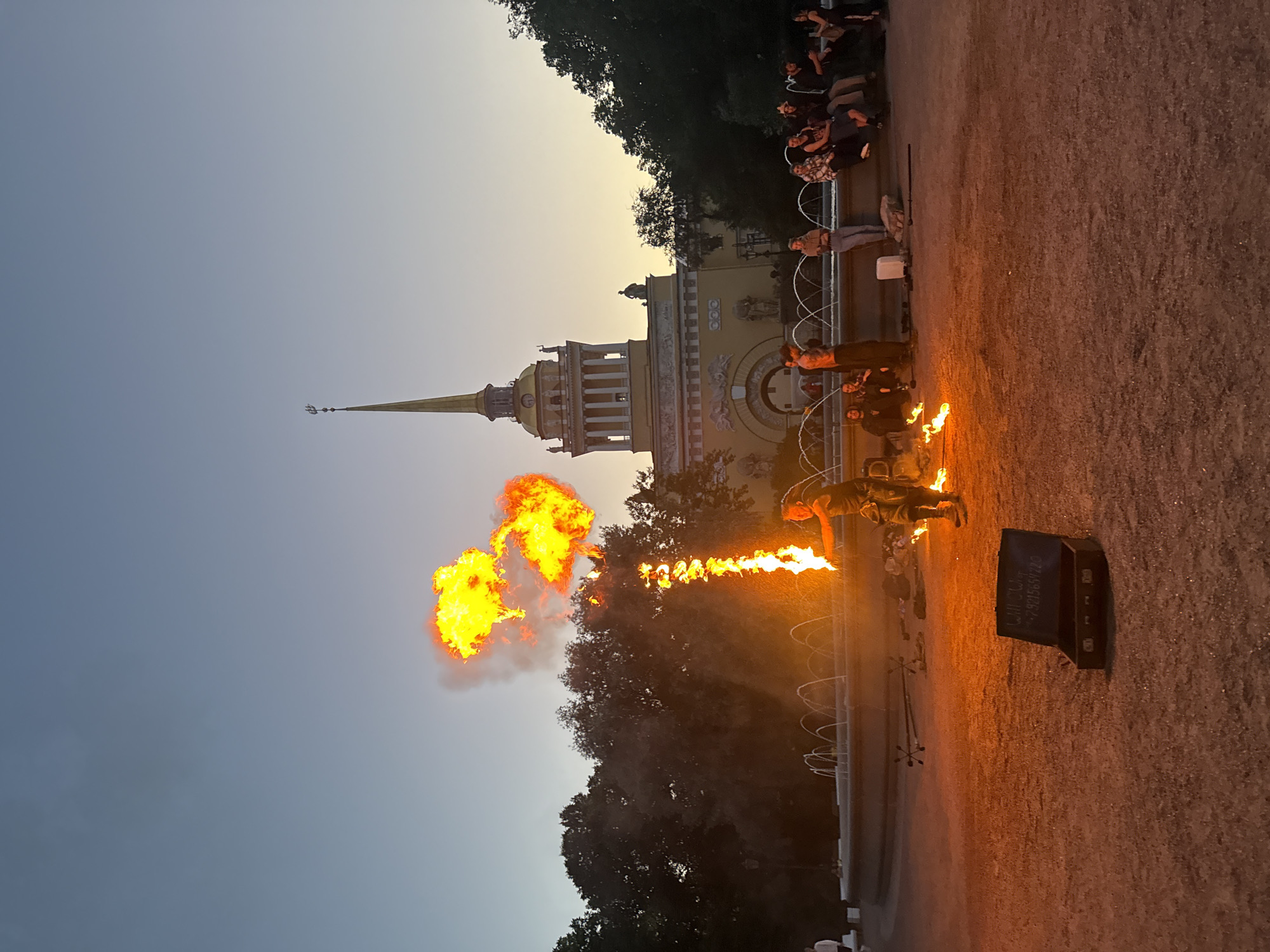 The image depicts a fire-breather performing in front of the Admiralty Building in Saint Petersburg, Russia, during twilight. The performer, a man wearing dark clothing, stands in the center of the frame, exhaling a large plume of fire that shoots upwards and to the left. He is holding a flaming object in his right hand. In front of him, on the ground, is an open black case with some writing on it. Scattered around the performer are fire-related props, including what appear to be staffs with four prongs.
Behind the performer, a fountain is visible, with water jets creating a misty effect. A crowd of onlookers is seated and standing along the edge of the fountain, watching the performance. To the left of the performer, a dark line of trees forms the background, and to the right, more trees are visible.
The Admiralty Building, a prominent landmark in Saint Petersburg, is in the background. It is a large, pale yellow building with classical architectural elements, topped by a tall, golden spire with a ship weathervane. The sky above is a gradient of blue and orange, indicating the setting sun. The ground in the foreground is covered in gravel. The overall atmosphere is that of an outdoor evening performance in a public space.
