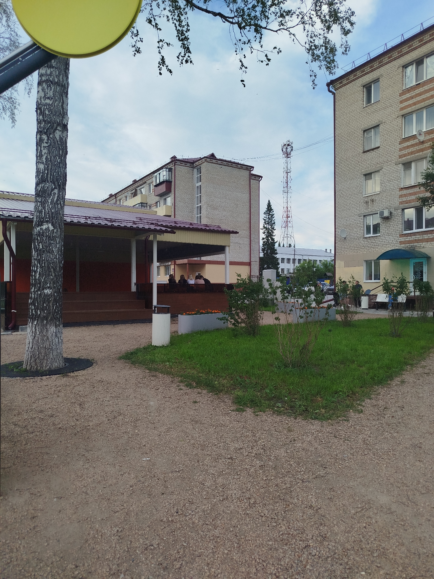 The image shows a view of an urban area in Zavodoukovsk, Russia, on a cloudy day. In the foreground, a large tree trunk with textured bark stands to the left, partially obscuring a yellow circular object above it. A gravel path extends into the middle ground, where a covered outdoor seating area with a red roof is visible. Several people are seated under the shelter. Beyond the seating area, a green lawn with bushes and flowers is present. Two multi-story brick apartment buildings, one light gray and the other with reddish-brown accents, are prominent in the background. A tall, lattice radio tower is visible between the buildings. The sky is overcast.