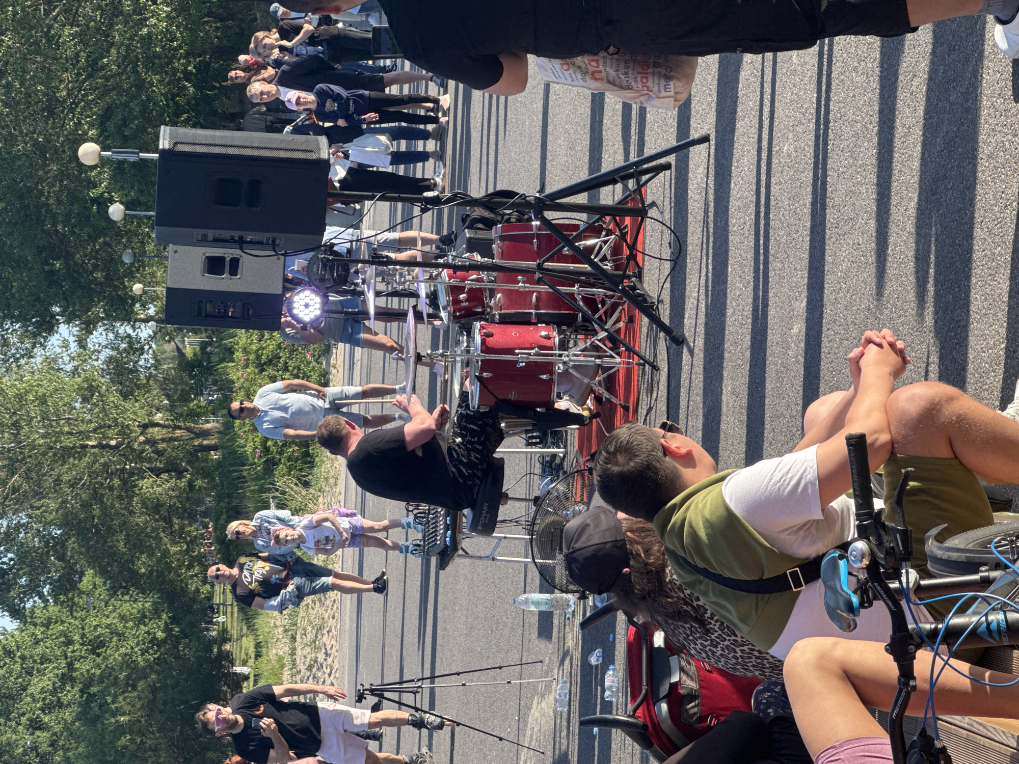 A group of people are gathered outdoors on a sunny day, near what appears to be a waterfront or park area in Tallinn, Estonia. In the foreground, a drummer is playing a red drum set. To the left of the drummer, a tripod is set up, possibly for recording the performance. In the background, several other individuals are standing and observing, some appearing to be tourists or locals enjoying the event. One individual in the background is holding a white bag with text on it. The environment includes trees and a paved surface, suggesting a public outdoor space.