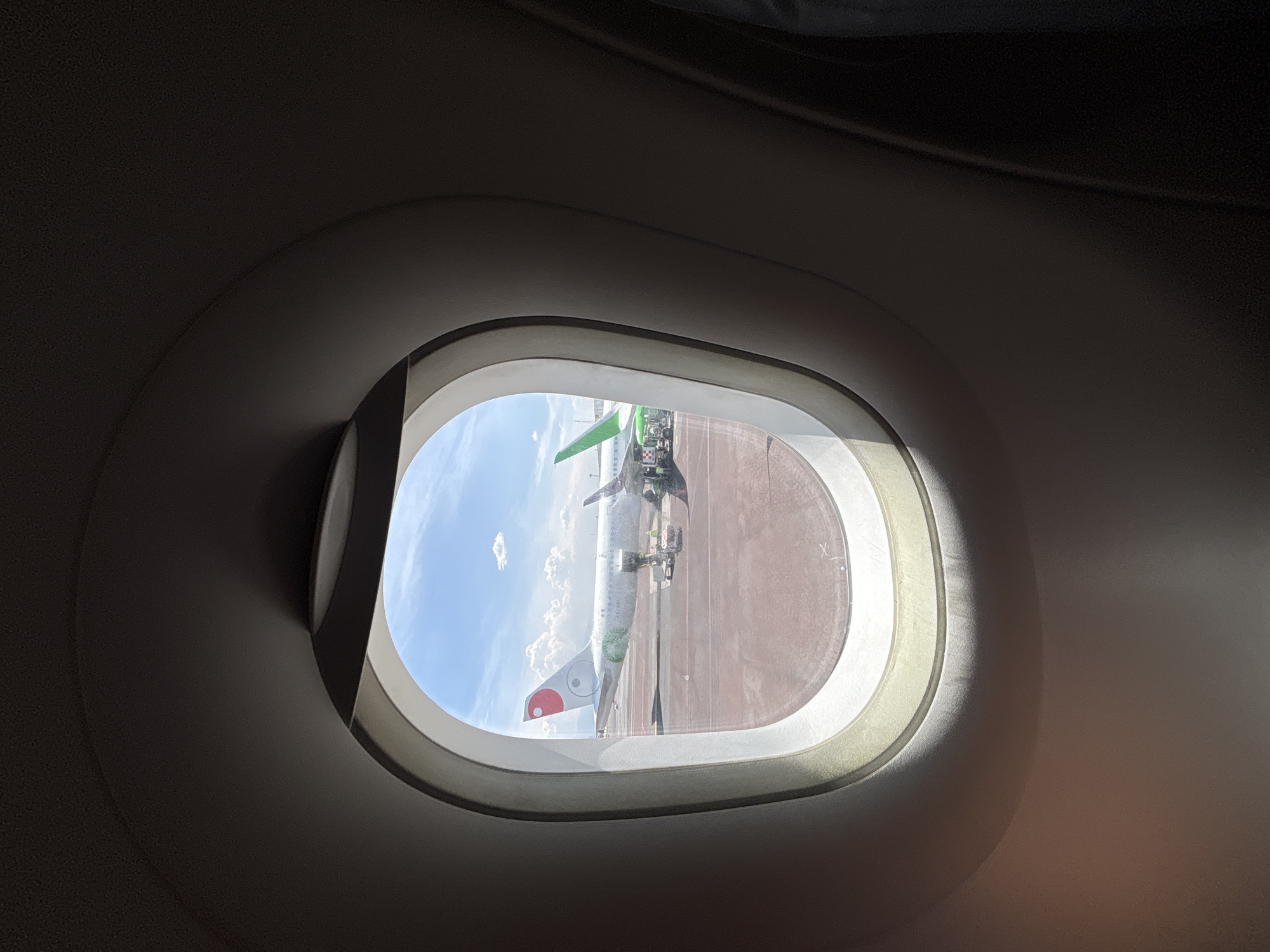 The image shows an airplane window with a view of two airplanes on the tarmac under a partly cloudy blue sky. The airplane on the left has a white fuselage with a red and white circular logo on its tail, resembling a Yin Yang symbol with an additional red circle. The airplane on the right has a green tail fin and a green design on its wing. Several figures, possibly ground crew, are visible near the airplanes, some wearing high-visibility vests. The tarmac appears to be reddish-brown. The interior of the airplane cabin is dark, with the window frame creating a distinct oval shape in the foreground. There is no visible indication of Mexico City.