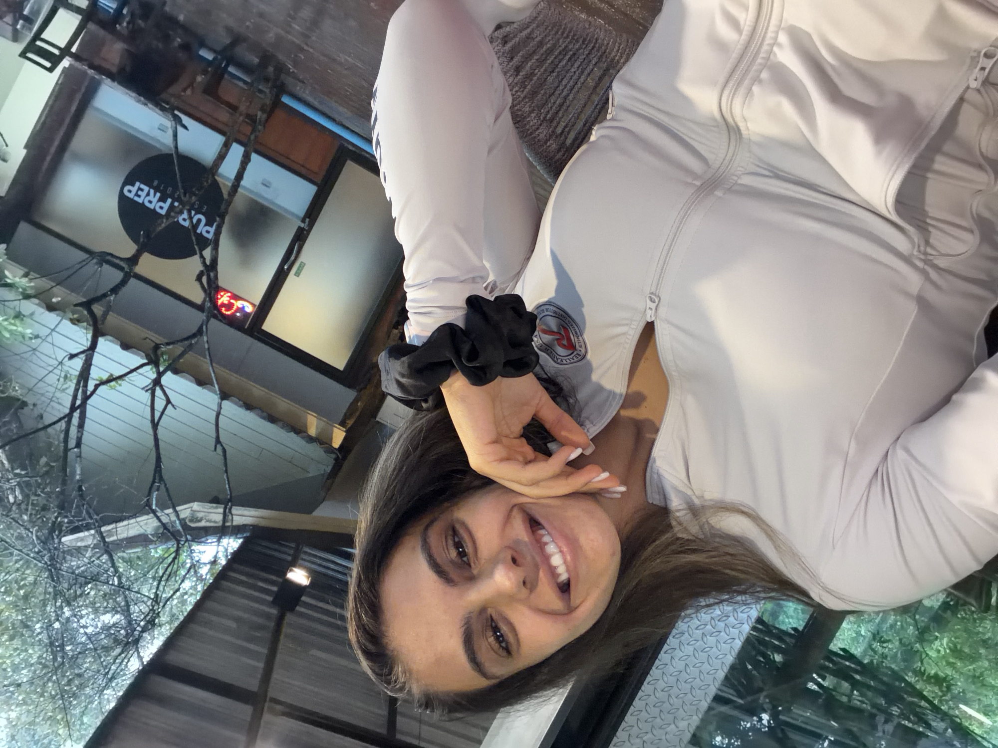 A young woman with long brown hair smiles and poses for a selfie. She wears a light-colored tracksuit with a visible patch on the left shoulder. Her left hand, adorned with a black scrunchie on her wrist, gently touches her cheek. Behind her, the "Pur Prep" sign is visible on the glass window. The location appears to be outdoors, possibly a patio or sidewalk area in Chalong, Thailand. The environment includes architectural elements and natural foliage.