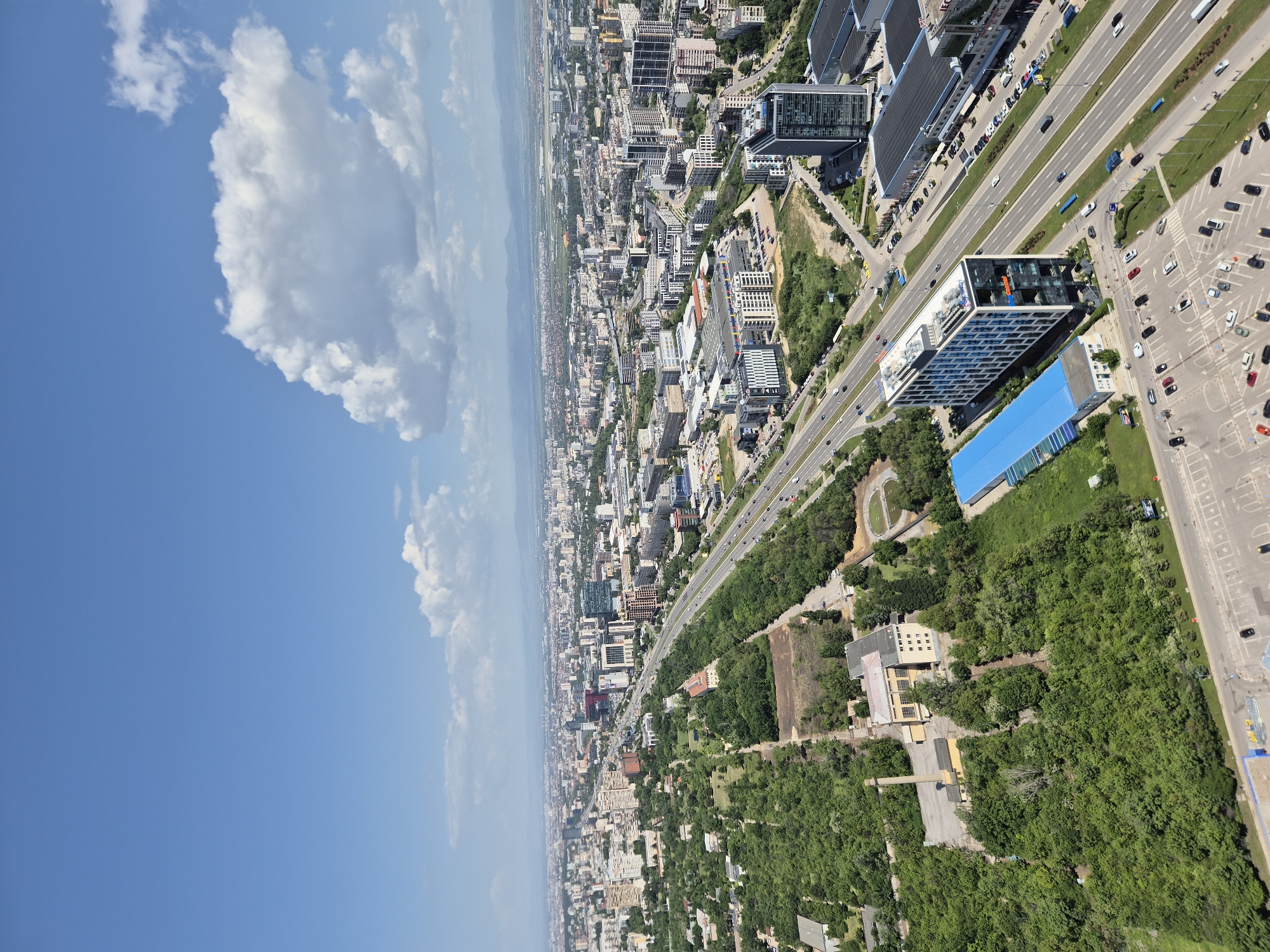 The image depicts an aerial view of Sofia, Bulgaria, characterized by a blend of urban development and natural landscape. In the foreground, a blue sky with scattered clouds dominates the upper-left portion of the frame. Progressing towards the right, the cityscape unfolds with a dense arrangement of buildings, ranging from low-rise residential structures to taller commercial buildings. A prominent highway runs through the center of the city, with vehicles visible in motion. Patches of greenery, including parks and wooded areas, are interspersed among the built environment. In the upper-right, there are parking lots with cars parked. Overall, the scene presents a juxtaposition of the city's infrastructure, architecture, and natural elements.