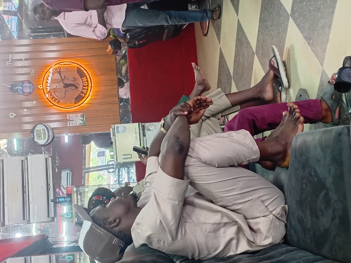 The image displays the interior of KAKNA HAIRCUTTING SALON in Dorayi, Nigeria. In the foreground, a dark-skinned male is seated on a dark grey upholstered sofa, facing right. He wears a light beige long-sleeved shirt, matching trousers, and a beige cap with a mesh back. His bare feet are visible, resting on the sofa, and he holds a dark-colored smartphone in his left hand, with his hands clasped over his knee. Partially visible behind him, another male is seated, wearing dark (possibly burgundy) trousers, with his bare feet resting on the checkered floor next to dark slippers. The floor is tiled with a black and white checkered pattern.
The background reveals a mirrored wall with two white split-unit air conditioners mounted high. A digital clock displaying "4:51" is visible on the wall. Centrally, a circular wooden sign with an orange LED backlight reads "KAKNA HAIRCUTTING SALON," featuring a graphic of the African continent and a barber's pole. To the right, a male stands, wearing a light pink long-sleeved shirt and blue jeans, with sandals on his feet. A red-painted partition or wall section is visible beneath the mirrors on the right side. Reflections of other individuals and salon items are observable in the mirrors.