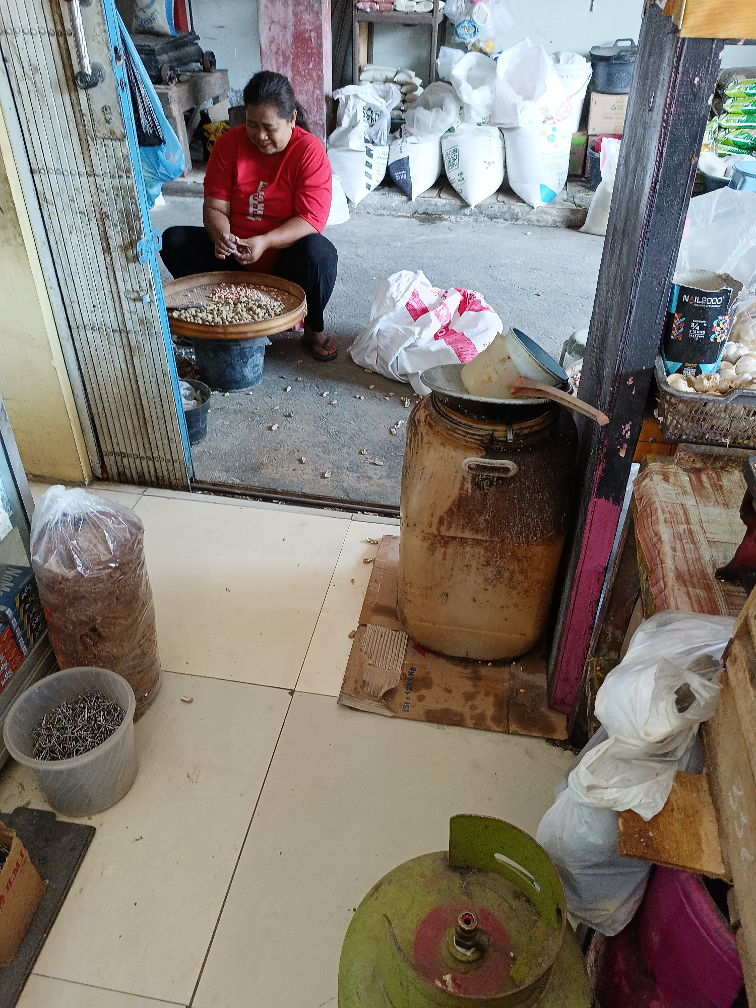 An Asian woman in a red t-shirt and black pants is sitting on the floor in front of a large metal tub filled with peanuts. She is sorting through the peanuts with her hands. The scene appears to be a market or a storage area, with various bags and containers in the background. There is a large plastic barrel, a bag of what appears to be rice, and a basket of garlic. The floor is tiled, and there are some scattered peanuts on the ground. The overall lighting is dim, suggesting an indoor or shaded outdoor environment. The woman is looking down at the peanuts, focused on her task. The image was taken in Manisrenggo, Indonesia.