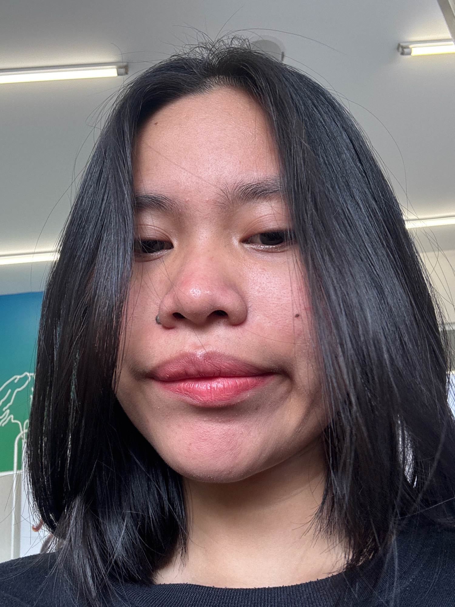 The image is a close-up selfie of a young Asian woman, likely taken indoors under fluorescent lighting. She has dark, straight hair parted in the middle and falling to her shoulders. Her facial expression is neutral to slightly amused, with her lips pursed. She is wearing a black crew-neck top. There is a subtle mole on her left cheek. The background shows a light-colored ceiling with fluorescent light fixtures, suggesting an indoor environment such as a classroom or office in East Jakarta, Indonesia. A blurred green and white poster is visible on the left side of the background.