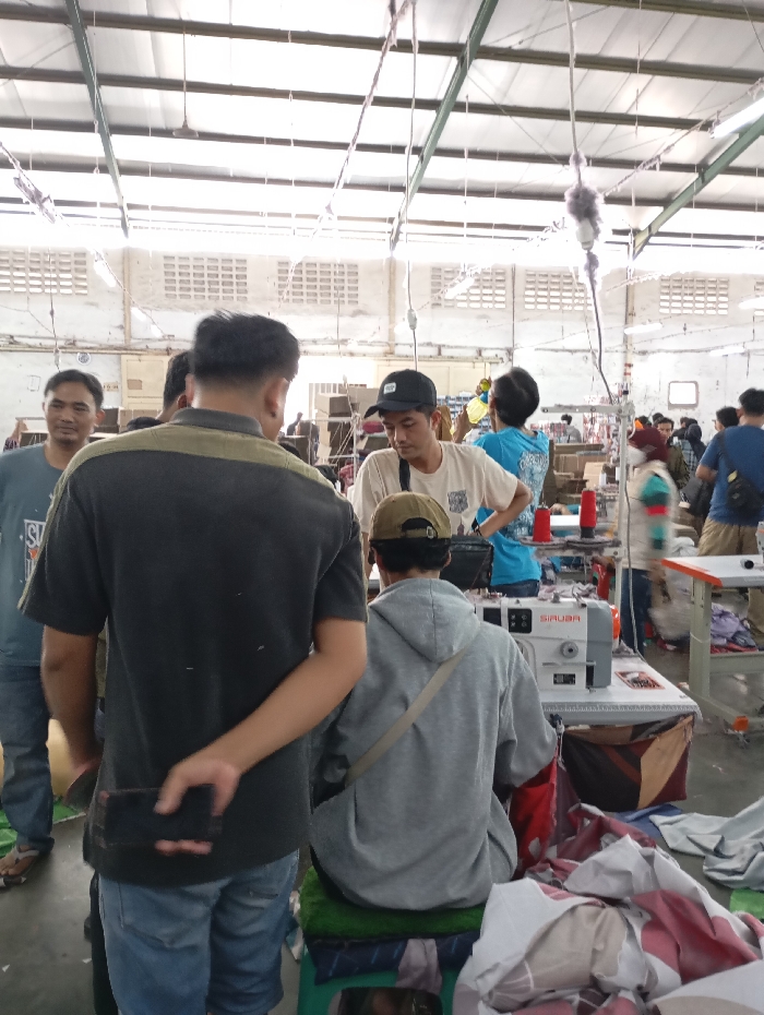 The image captures an active indoor scene in a garment factory or workshop located in Katapang, Indonesia, likely during the daytime. The setting features a high ceiling with exposed metal support beams and corrugated roofing, along with white walls containing multiple ventilation blocks, typical of an industrial building. Wires, cables, and fluorescent lighting fixtures hang from the ceiling.
Numerous individuals, mostly men, are present. In the foreground, a man with his back to the camera, wearing a dark grey t-shirt and blue jeans, holds a black phone behind his back with his right hand. To his left, another man in a blue t-shirt smiles faintly, looking towards the camera. To the right, a man in a light grey hoodie and a cap is seated on a green stool, facing away from the camera, operating a white industrial sewing machine with the brand "SIRUBA" visible on its side. Red and blue spools of thread are on the machine table. Piles of fabric scraps and textile products are scattered on the table next to the sewing machine and on the floor.
Other workers are visible in the mid-ground and background, including a man in a white t-shirt and black cap looking downwards, and another in a blue t-shirt further back. People appear to be engaged in various tasks related to textile production, standing, observing, or actively working at machines. The overall atmosphere suggests a busy, productive environment. No strong emotions are overtly displayed, but the individuals appear focused on their activities.