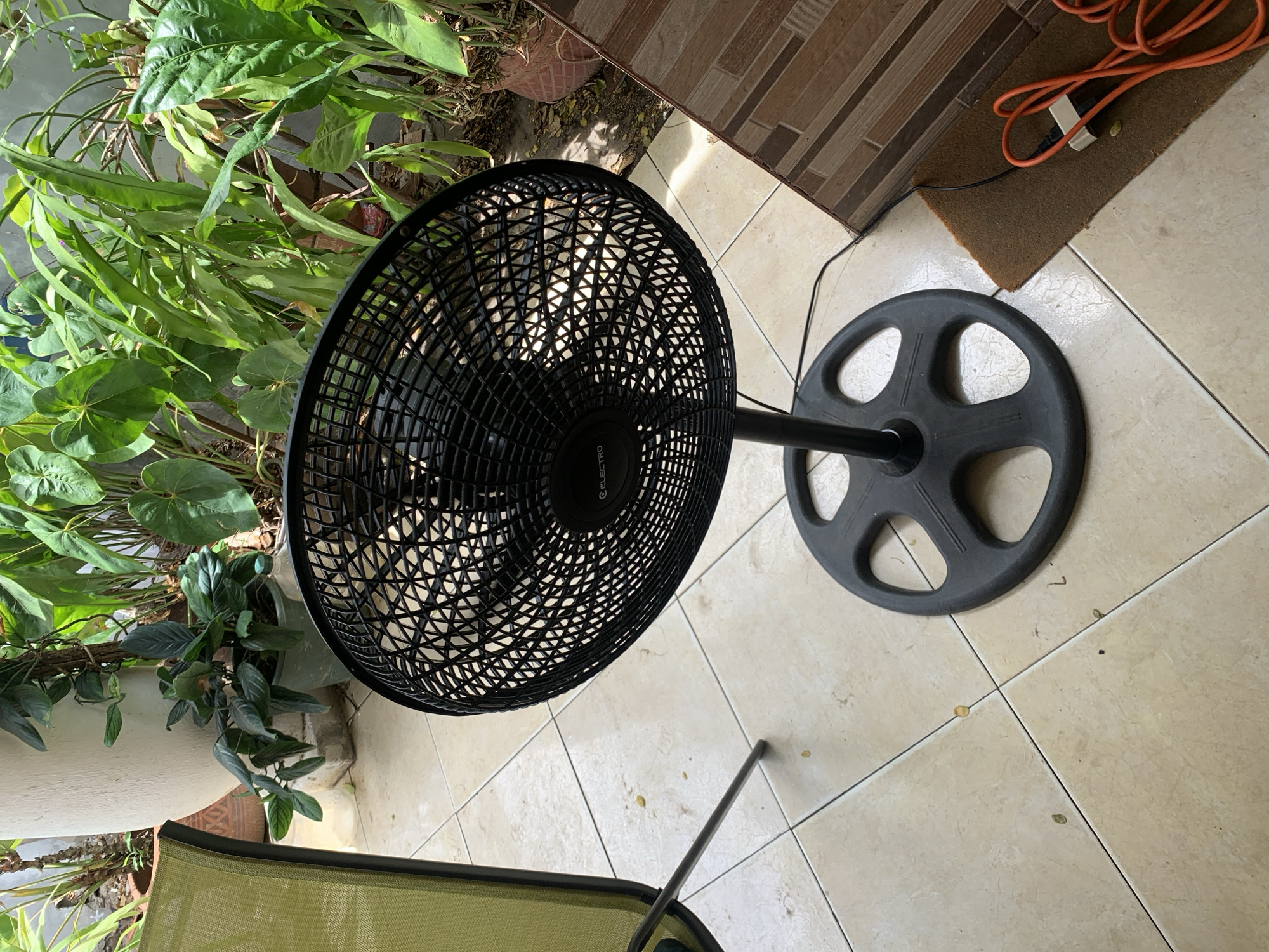 The scene depicts an outdoor or semi-outdoor patio area in Santa Ana, Venezuela, under natural daylight. A black "ELECTRO" brand stand fan is centrally positioned, featuring a circular mesh guard, a black pole, and a multi-lobed base. Its power cable extends along the light-colored tiled floor towards the right. A partially visible chair with a lime green fabric backrest and black metal frame is located on the left. The tiled floor exhibits dark grout lines and is scattered with small pieces of debris. In the background, a dense array of green foliage from various potted plants, including a tall white planter and other ceramic pots, forms a natural backdrop. On the right side, a wall or low structure with horizontal brown rectangular tiles is visible. A small, dark brown fibrous mat rests against this tiled surface, holding a coiled orange electrical cable with a white adapter. No individuals are present in the image.