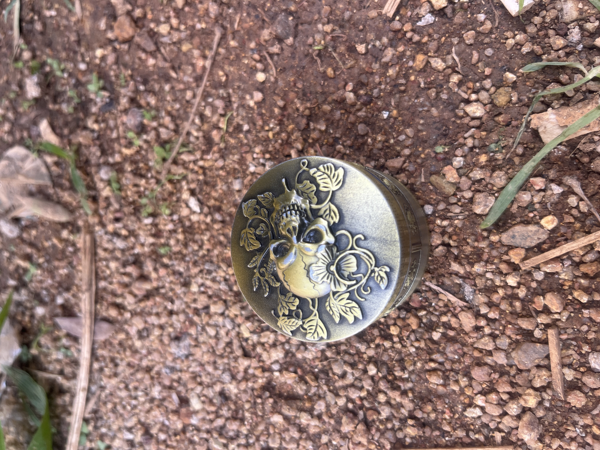 The scene is located in Katon Rikkos, Nigeria. The image depicts a small, ornate, cylindrical metal container resting on dry, reddish-brown soil densely mixed with small, angular gravel pieces. The container, appearing to be made of aged brass or bronze, features a decorative lid with a raised design. This design prominently displays a stylized human skull positioned centrally, encircled by intricately swirling floral patterns and leaves. Sparse patches of green foliage and scattered dried organic debris, including small twigs, are visible across the soil surface in the foreground and background. No individuals, animals, or active interactions are present.